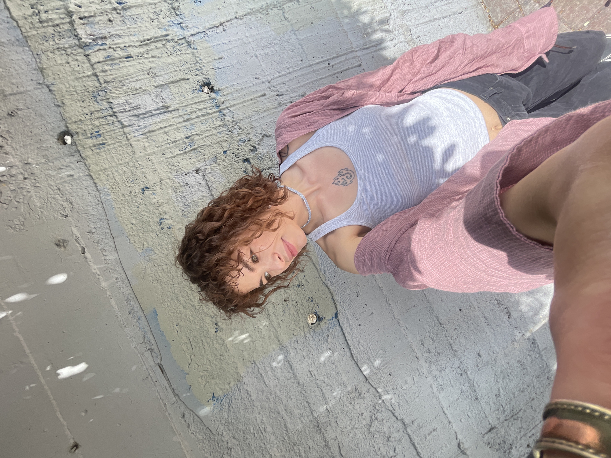 The image shows a person lying on a concrete surface, possibly a wall or a slab, with the camera positioned above. The person has curly reddish-brown hair and is wearing a white tank top, dark gray pants, a light pink long-sleeved shirt, and a silver necklace. A tattoo is visible on the person's left shoulder. The concrete surface has a rough texture with some discoloration. Sunlight creates patterns of light and shadow across the scene. The location might be Rafailovići, Montenegro, based on the architecture and environment.