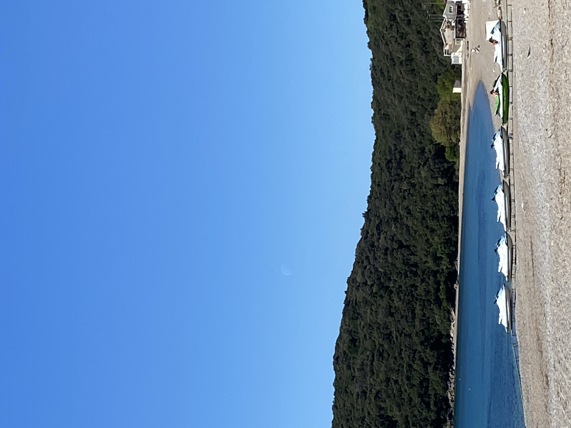 The image shows a coastal scene in Prijevor, Montenegro. The view is rotated 90 degrees counterclockwise. The sky, a clear blue, occupies the left portion of the image. A dense, green forest line extends along the right-center. Along the right edge is a beach with beige sand. Light blue water lies adjacent to the beach, punctuated by what appear to be white structures (possibly umbrellas or canopies). A few buildings are visible at the top right. A faint circular form is observable in the sky, possibly the moon.