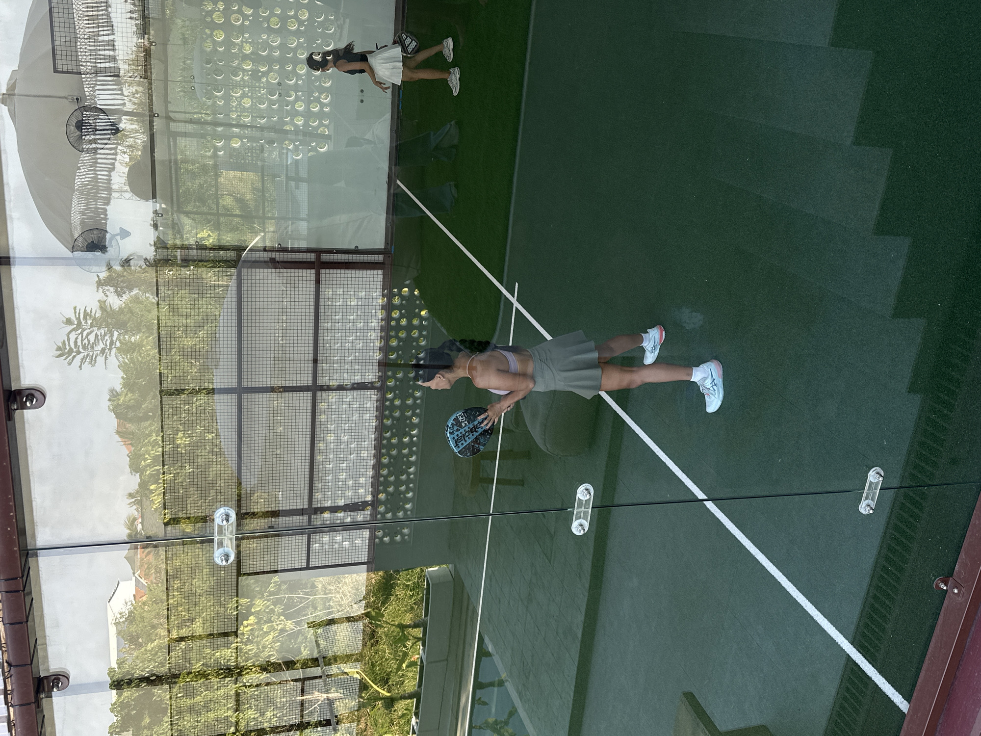 A woman wearing a light purple sports bra and a green skirt stands on a green court with white lines, holding a blue and black paddle. She is looking up at the paddle in her hands. Her light blue and white sneakers are visible. In the background, another woman in a black top and white skirt is walking away, also holding a paddle. She is wearing white sneakers and headphones. The court appears to be an outdoor padel court enclosed by glass walls. Through the glass, trees, fences, and buildings are visible. There is a large umbrella with two fans attached in the background, along with a wall decorated with circular patterns. The scene is well-lit, suggesting daytime. The court has a synthetic turf surface.