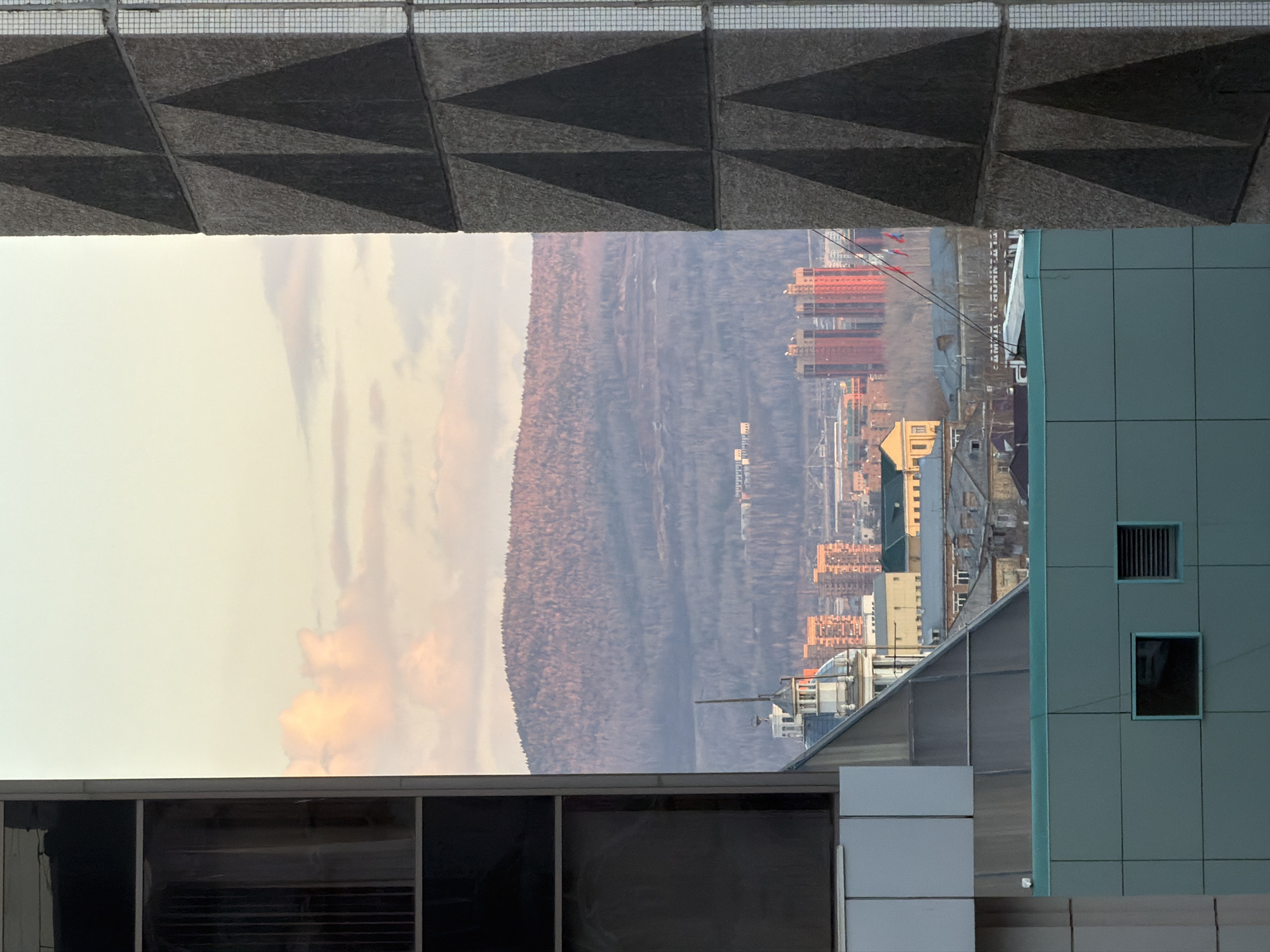 The image, when viewed upright, displays an expansive urban and natural landscape, likely captured from a high vantage point within a building. The scene is horizontally framed by a prominent architectural element in the midground.
In the immediate foreground (bottom), parts of two modern buildings are visible. On the left, a section of a facade with large, dark, reflective windows mirrors the sky. To the right, a light blue-grey paneled building features two small windows, one fitted with a grille, and an angled glass or metal roof structure extends from it.
Stretching horizontally across the middle of the frame is a significant concrete structure, possibly an elevated walkway, bridge, or part of a building's underside. It showcases a distinctive geometric pattern of dark and light triangular facets. A fine metal grid or railing runs along its upper edge.
Beyond this concrete frame, the background reveals a dense cityscape with numerous multi-story buildings, many exhibiting reddish or orange facades, characteristic of residential blocks. Several construction cranes rise above the urban fabric. Faint power lines or communication cables can be seen traversing the middle distance.
The far horizon is marked by a long, undulating range of hills or mountains, densely covered in what appear to be dormant or leafless trees, creating a muted brownish-grey texture.
The sky, filling the top portion of the image, is light and hazy, adorned with soft, warm-toned clouds exhibiting yellowish-pink hues. This suggests either an early morning sunrise or a late afternoon sunset. No specific activities or individuals are visible, and there is no discernible text. The scene depicts a city, consistent with Krasnoyarsk, Russia, situated against a backdrop of natural mountainous terrain, viewed through a distinctive architectural aperture.