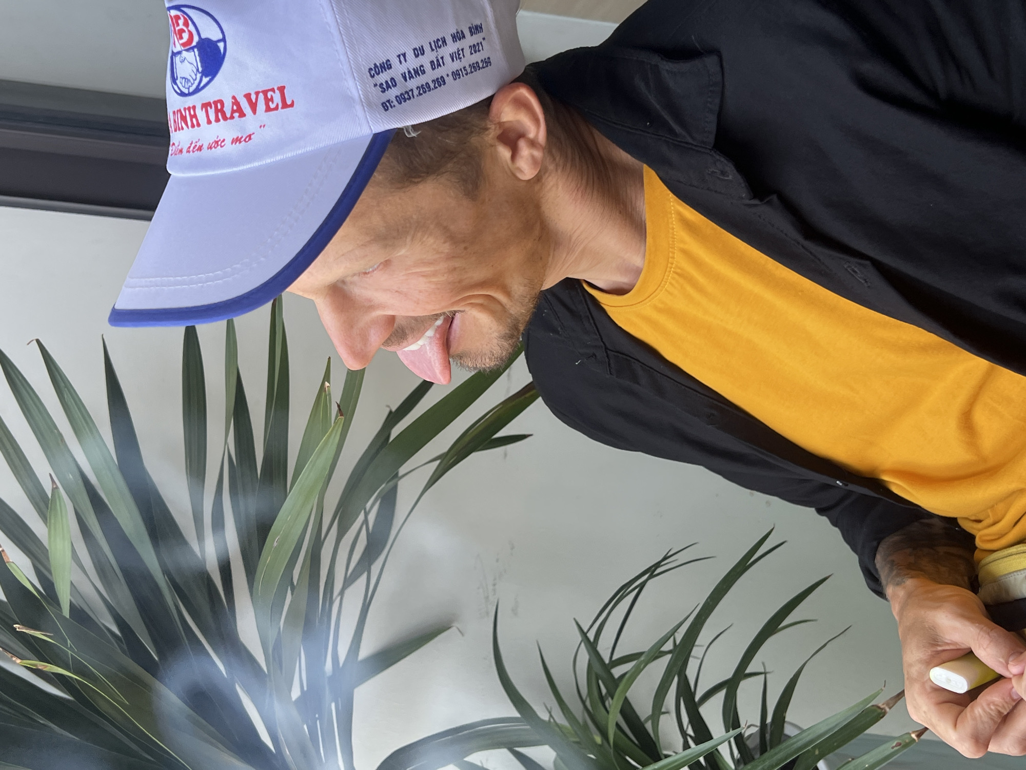 A man wearing a white baseball cap, a black jacket, and a yellow t-shirt is sticking his tongue out in front of a plant. The cap is printed with "BINH TRAVEL" and "Đểm đến ước mơ" as well as "CÔNG TY DU LỊCH HÒA BÌNH", “SAO VÀNG ĐẤT VIỆT 2021", and phone numbers. He is holding a vape pen. There is smoke emanating from the plant. The city of Nam Natural Heritage Area Trang, Vietnam is not visible.