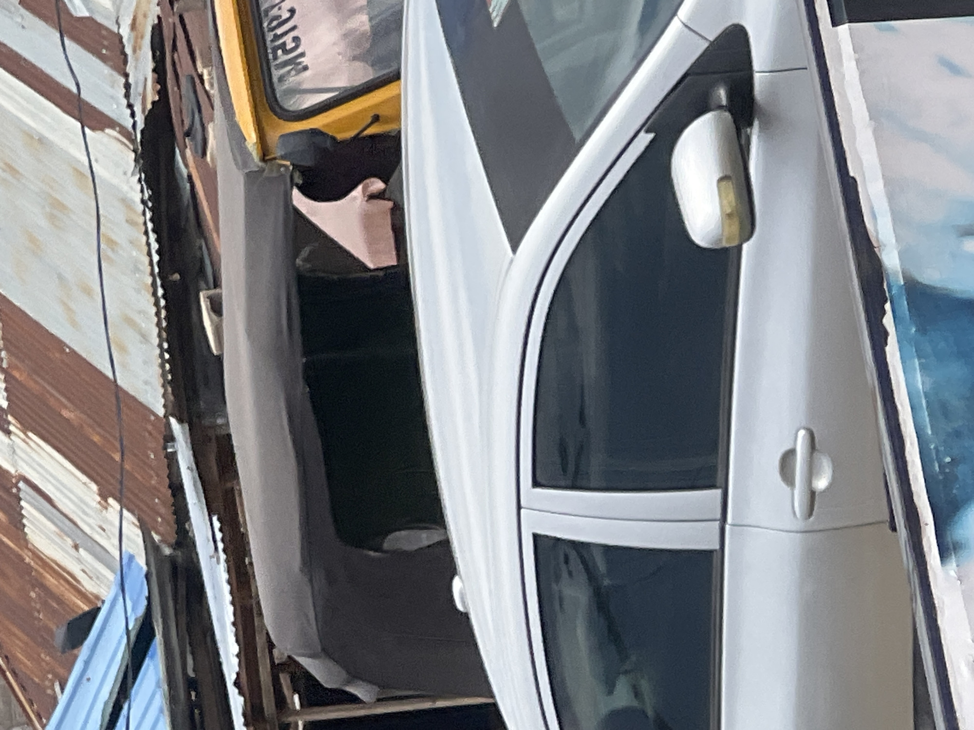 The image captures a street scene in Jalingo, Nigeria. In the foreground, a silver sedan is visible, with its side door handle and side mirror in focus. The car appears to be parked or in slow motion. Behind the sedan, a yellow auto-rickshaw (Keke Napep) is partially visible, its roof and part of its window with text reading "METRO" are discernible. Further in the background, a makeshift structure with a corrugated iron roof, showing signs of rust, is present. The overall environment suggests an urban setting with a mix of vehicles and informal structures.