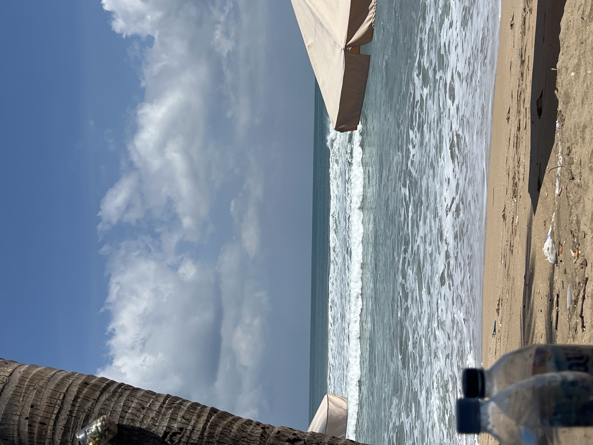 The image depicts a beach scene in Kuta, Indonesia. In the foreground, two plastic water bottles with blue and black caps are visible on the sand. The beach is sandy and strewn with debris, including plastic wrappers and sticks. In the middle ground, the ocean is visible with rolling waves and white foam. Two beach umbrellas are positioned on the right side of the frame, one partially visible and the other more prominently in the foreground. A palm tree trunk is on the left side of the image, with a glass jar attached to it containing some items. The sky is blue with scattered white clouds. The overall scene suggests a typical beach environment, potentially impacted by litter.