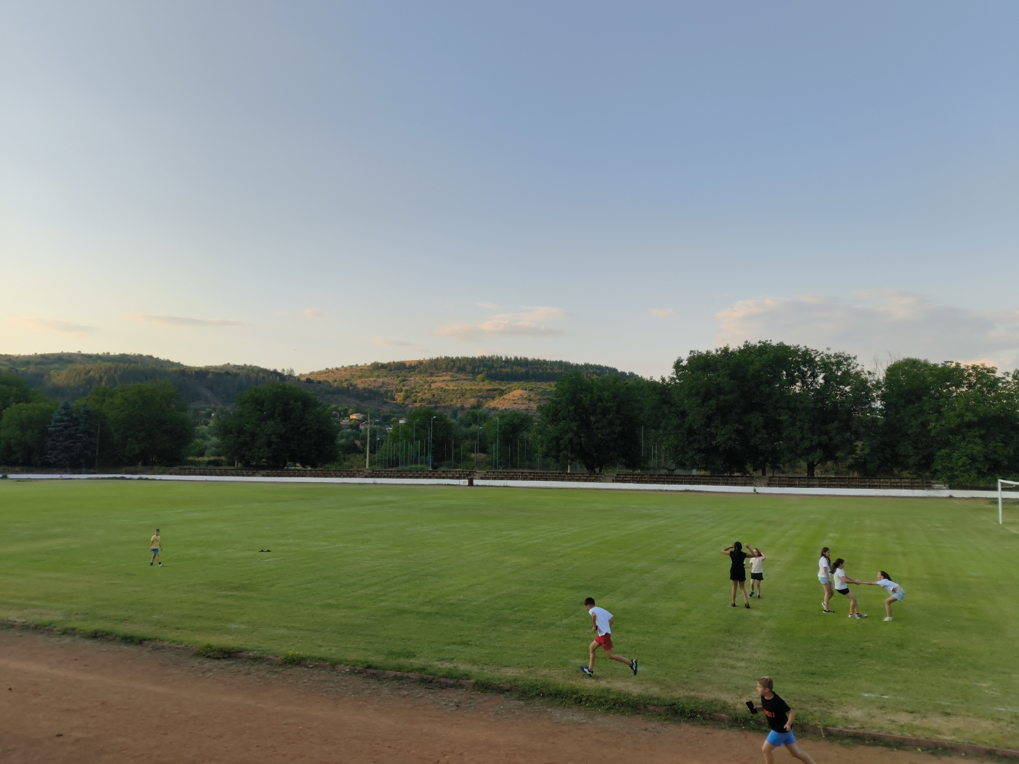 The image shows an outdoor athletic field with a group of people engaged in various activities. In the foreground, a dirt track runs along the bottom of the frame. The main field is a large expanse of green grass.
Several individuals are present on the field. On the far left, a lone person in a yellow t-shirt and blue shorts stands near the edge of the field. Further to the right, a person in a white t-shirt and red shorts is running. In the middle of the field, there is a group of people. Two individuals are interacting, possibly in a game or practice, with one holding the other's hand. Other individuals in this group are standing or appear to be moving. In the lower right corner, another person is running.
The background features a line of trees, with hills and more trees visible in the distance under a clear sky with some faint clouds. A white goalpost is visible on the far right side of the field. The lighting suggests it is late afternoon or early evening. This scene is consistent with the Boboshevo Stadium, located in Boboshevo, Bulgaria.