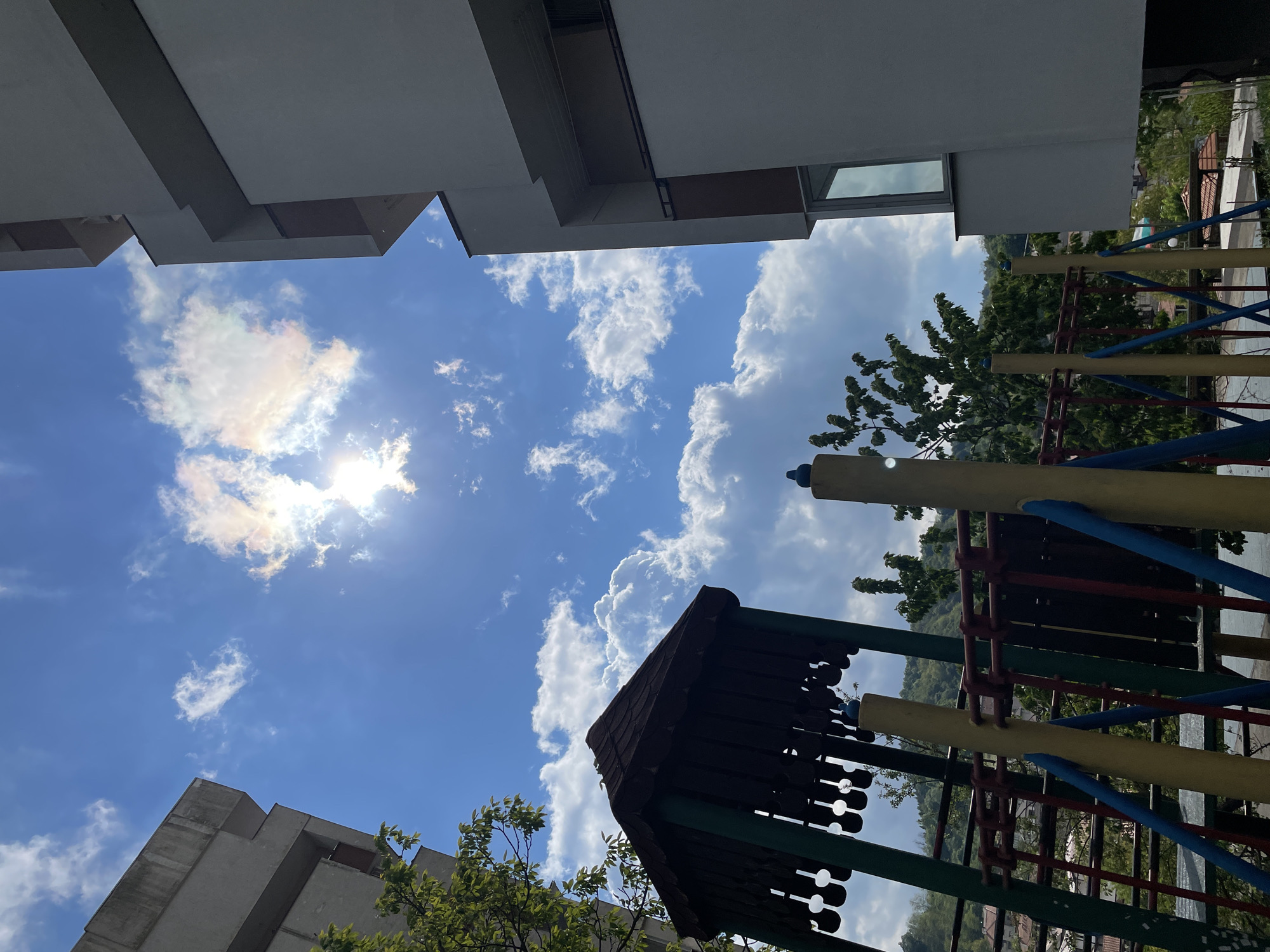 The image is an upward-angled view of a playground in the city of Zlatograd, Bulgaria. Buildings with angular, white facades are visible at the top and bottom of the frame, partially obscuring a bright blue sky with scattered white clouds. The sun is shining brightly, with some haze around it. A playground structure with a brown roof and colorful supports (yellow, blue, red, green) dominates the right side of the image. Green trees are interspersed between the buildings and the playground equipment. The camera is looking up.