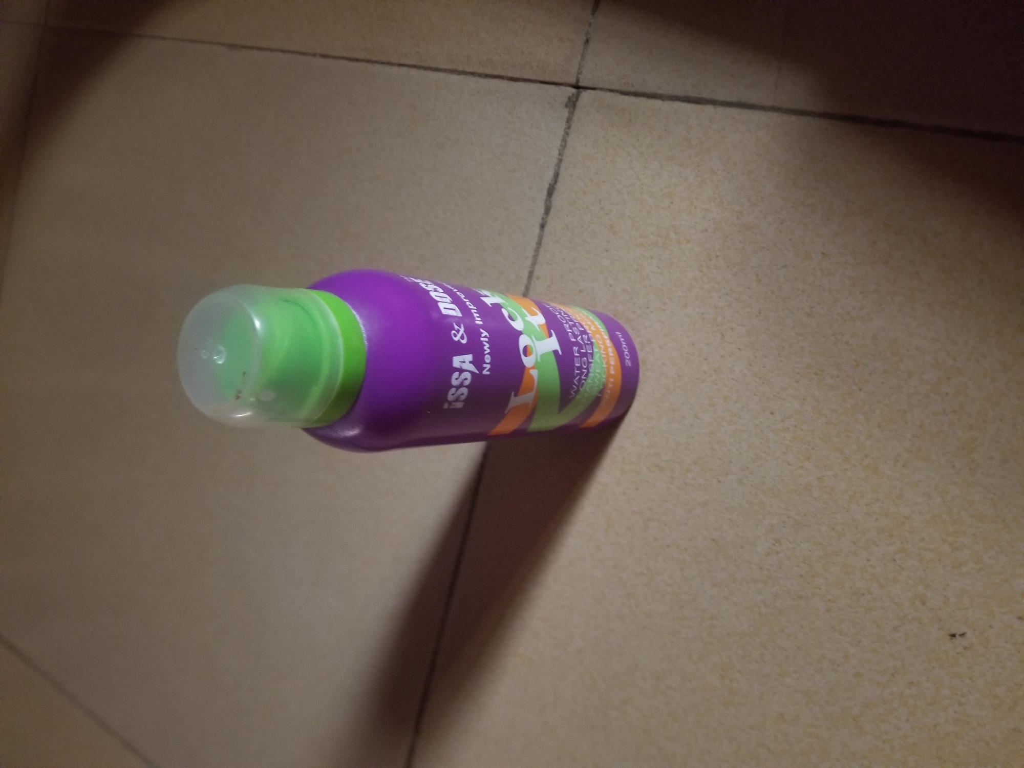 A purple bottle with a green cap sits on a tiled floor, casting a shadow to its left. The bottle has text on it that reads "issa & DOS", "Newly Improve", and "LOCK IT". Below this, smaller text indicates "WATER PROOF", "LONG LASTING", and "NON TRANSFER" with additional text that is unreadable. At the bottom of the bottle, "200ml" is visible. The tiled floor is light brown with dark grout lines. The lighting suggests an indoor setting. There are no people or animals present in the image. The scene is devoid of any specific environmental context that would indicate a location within the city of Jauro Yinu, Nigeria.