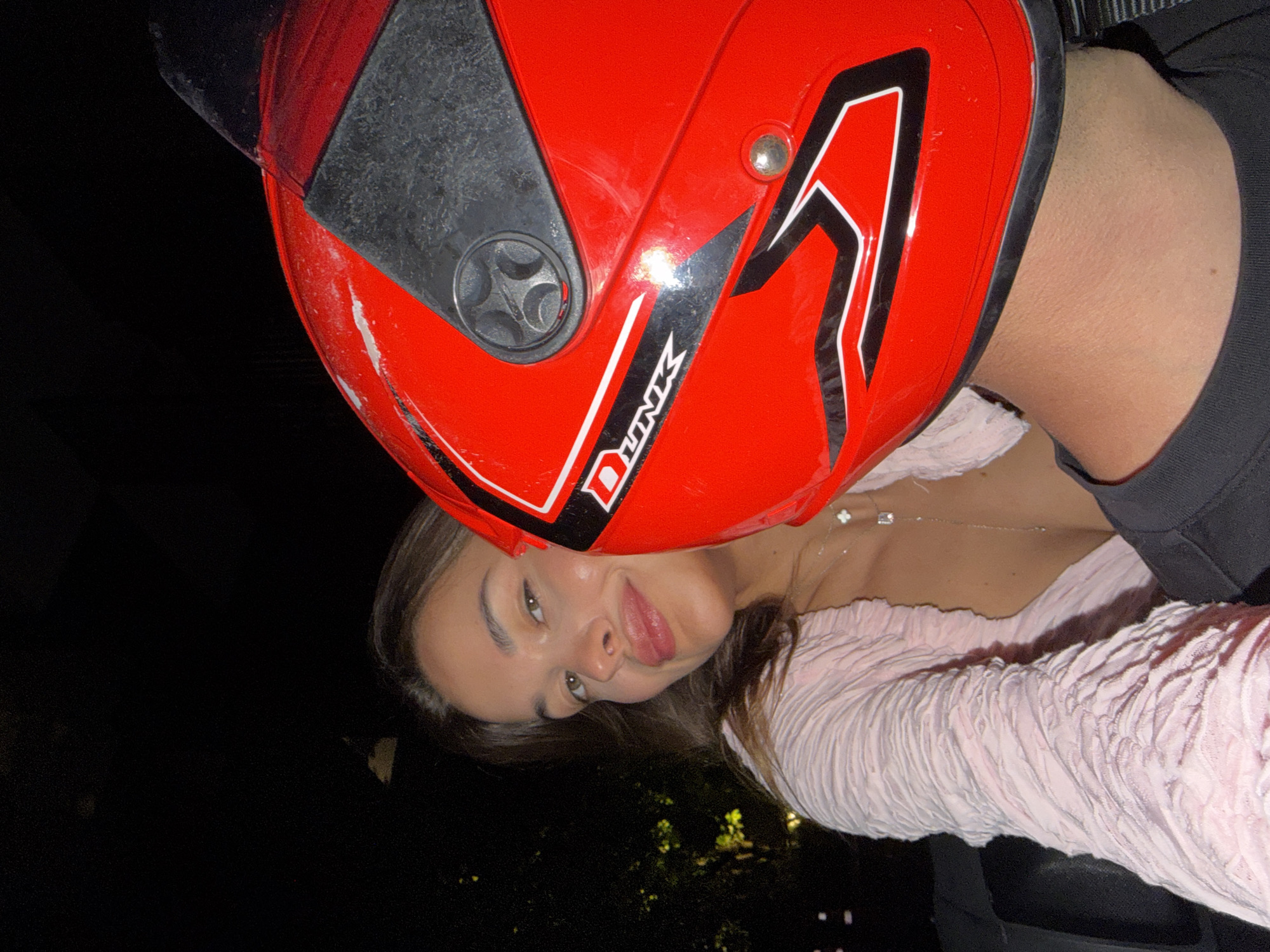A photograph depicts a person wearing a red motorcycle helmet with black and white accents, partially obscuring the top of their head. The person is wearing a light pink, textured top and a black garment near the neck. A necklace with a pendant is visible on their chest. The background appears dark, possibly indicating nighttime. There is insufficient information to determine if the photograph was taken in Choeng Thale, Thailand.