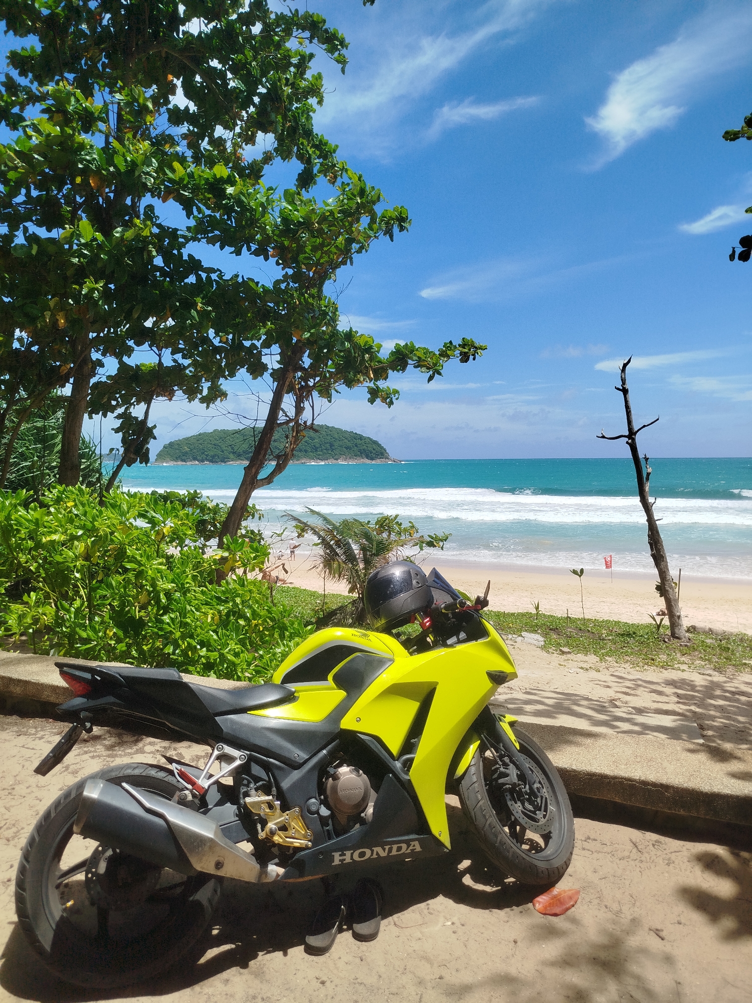 A bright yellow Honda motorcycle is parked on a sandy surface next to a concrete barrier. The motorcycle faces left in the frame, with its front wheel closer to the viewer. A black helmet rests on the handlebars. Behind the motorcycle, a beach stretches out with gentle waves breaking on the shore. In the distance, a small, tree-covered island is visible in the turquoise ocean under a partly cloudy blue sky. Lush green foliage and trees frame the scene on the left and in the foreground. A dried tree trunk stands on the beach to the right of the motorcycle. A red flag is planted in the sand near the water. The overall impression is a sunny day at a tropical beach.