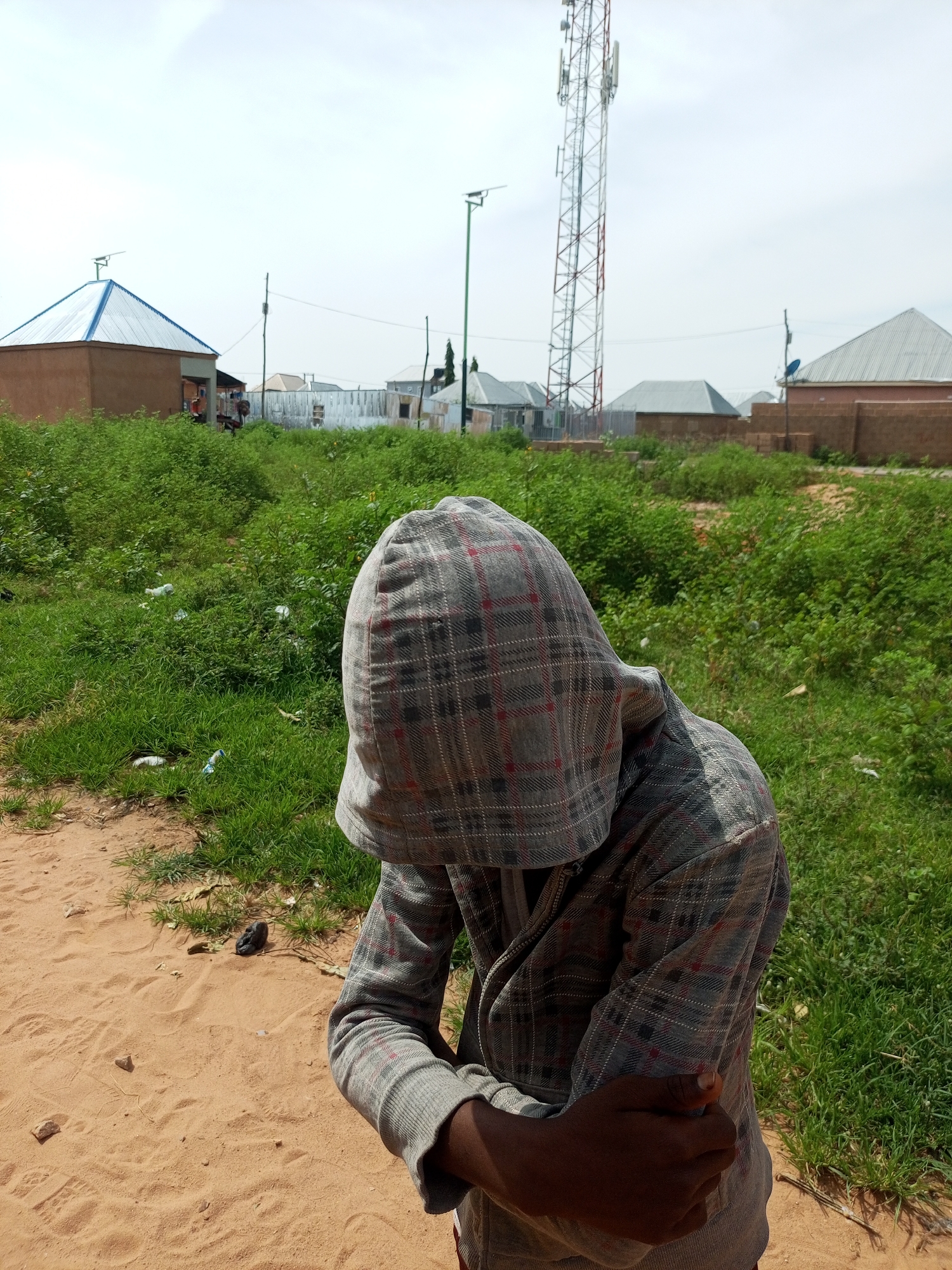 In Tempure, Nigeria, a young person wearing a hooded plaid jacket is positioned in the foreground, facing away from the camera and obscuring their face. Their left arm is bent and crossed across their torso, with their hand holding their right upper arm. The individual stands on a dirt path, with abundant green grass and scattered white debris to their right. In the background, several buildings with corrugated metal roofs are visible, as well as a tall telecommunications tower. The sky is overcast.