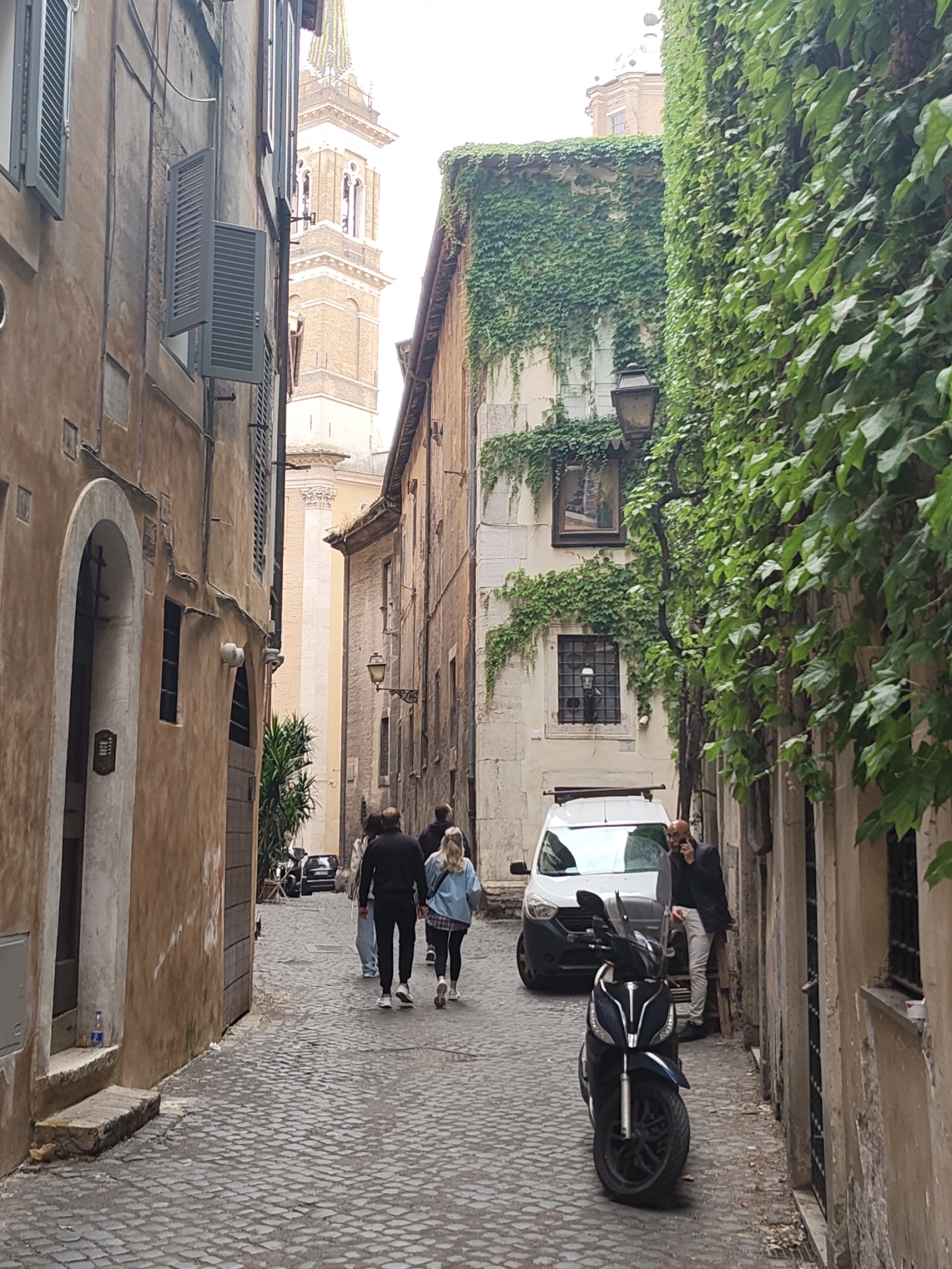 An eye-level, medium shot captures a narrow street in Rome, Italy, characterized by cobblestone paving. On the left, the architecture is marked by aged, multi-story buildings in earth tones, including an arched doorway and closed shutters. The right side features a building partially covered in thick green ivy. Three people are walking away down the street. Parked vehicles, including a white van and a black scooter, line the street, suggesting a limited space for pedestrians. A man stands near the van, engrossed in a cell phone conversation. In the background, a light-colored tower is visible, adding to the city's architectural landscape. The overall atmosphere conveys the charm and narrowness of a typical Roman street.