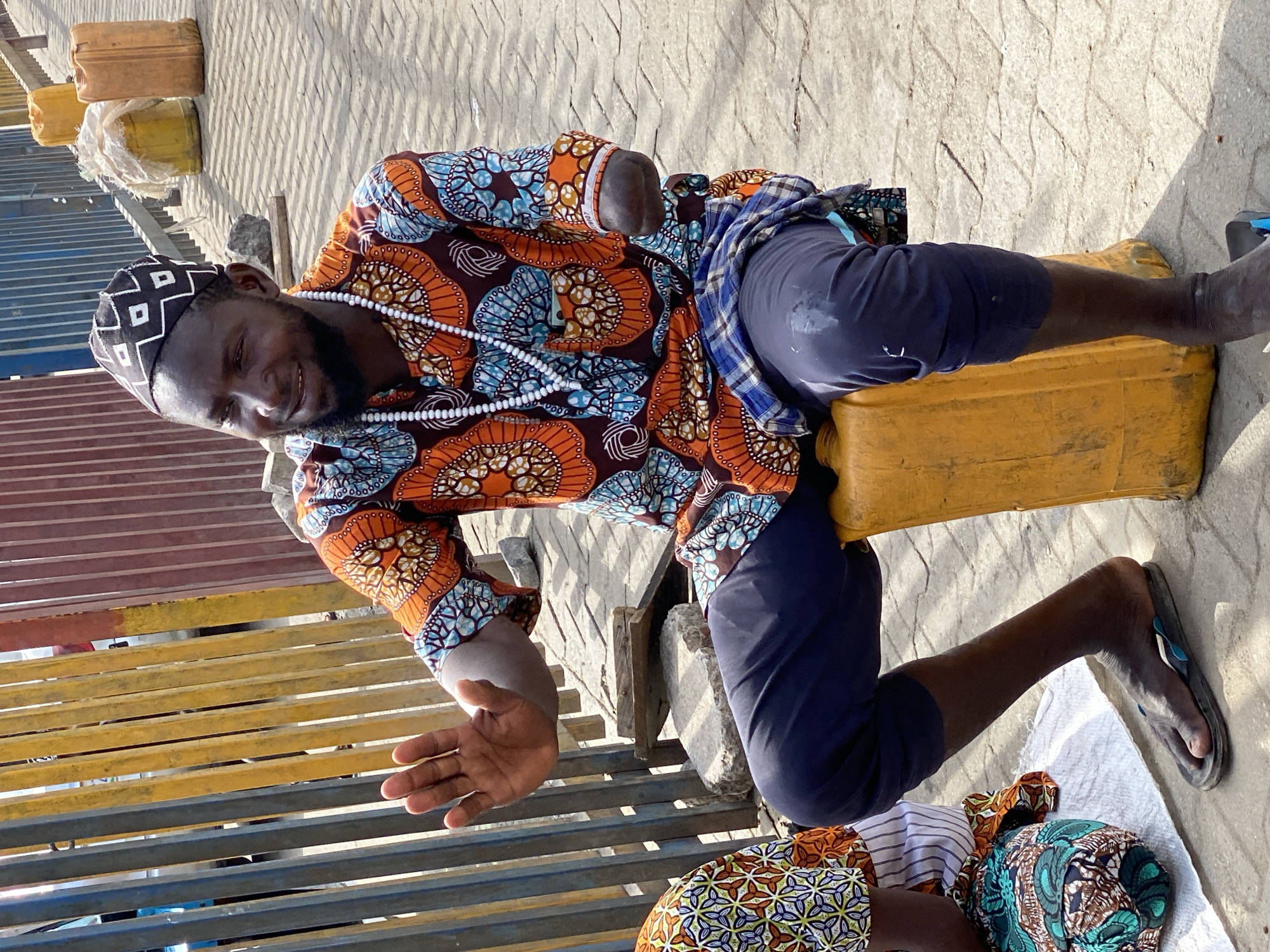 A man is kneeling on the ground next to a metal railing painted with yellow and blue stripes. He is leaning forward, his right hand outstretched. He is wearing a colorful patterned shirt, dark blue pants, a black and white patterned cap, and a white beaded necklace. He is kneeling on a yellow plastic container and has a blue plaid shirt tied around his waist. He is wearing a single flip-flop on his right foot. The ground is paved with interlocking concrete pavers. Yellow plastic containers are visible in the background. The person is sitting in Lekki, Nigeria.