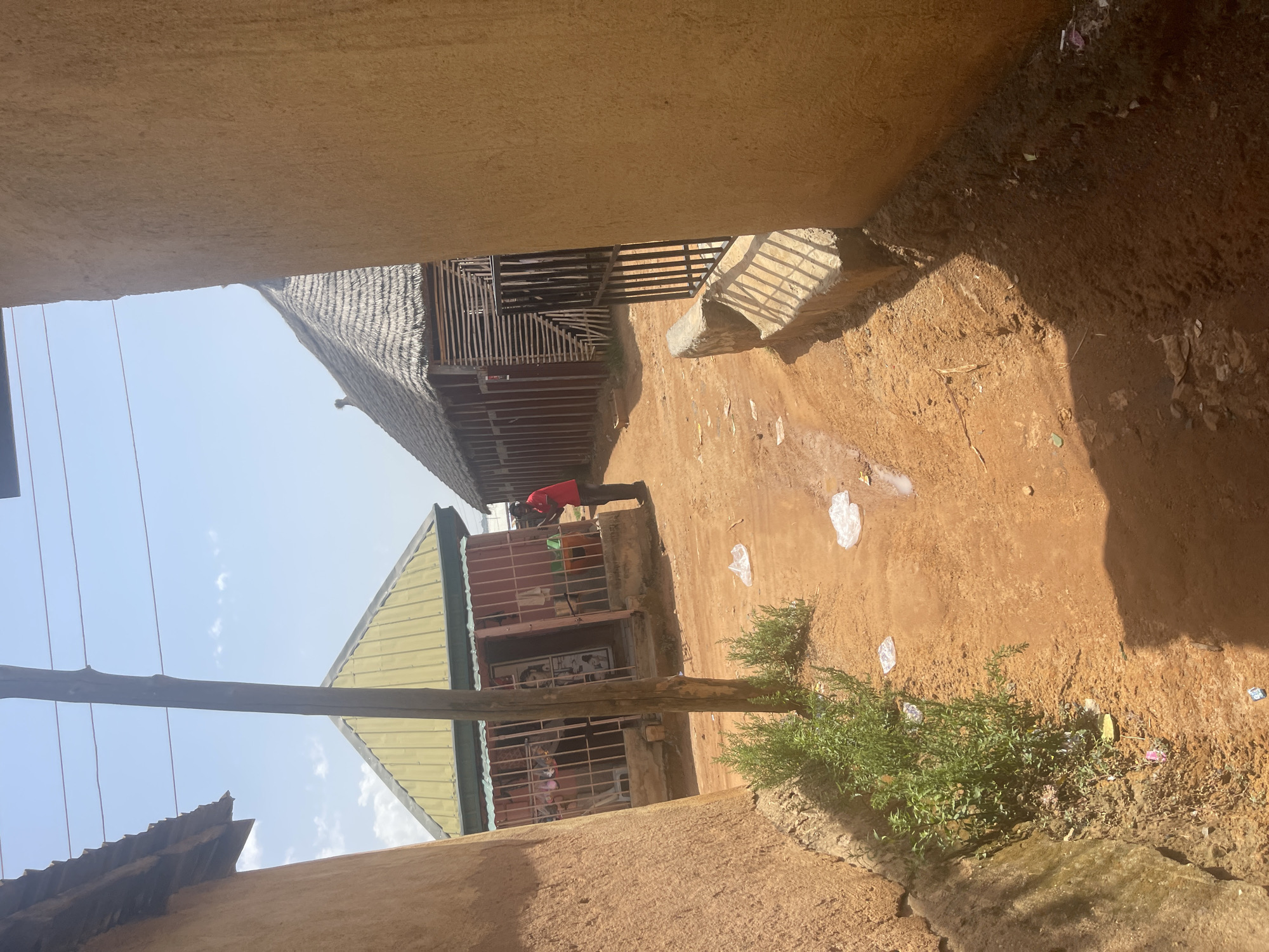 The image depicts an outdoor scene in Rafin Par, Nigeria. The perspective is from a low vantage point, looking up at buildings and the sky. The overall impression is of a somewhat rustic and possibly undeveloped area.
In the foreground, a rough, earthen ground with scattered debris, including plastic wrappers and some vegetation, is visible. To the left and right, earthen walls or structures rise up, framing the view. A wooden pole runs horizontally across the middle of the frame, obscuring part of the scene.
Beyond the foreground, two buildings are partially visible. The building on the left has a thatched or natural material roof and wooden slat walls, suggesting traditional construction. Adjacent to it is another building with a metallic, light-colored roof, possibly corrugated metal. This building appears to have barred windows and a red-colored exterior. A person wearing a red shirt is seen bending down near this building.
The sky is a clear blue, indicating it is daytime. The sunlight creates shadows, suggesting the sun is at an angle, typical of mid-morning or afternoon. The presence of the buildings, the type of construction, and the general appearance of the ground suggest a rural or semi-urban setting in Nigeria. There is no visible text in the image.
