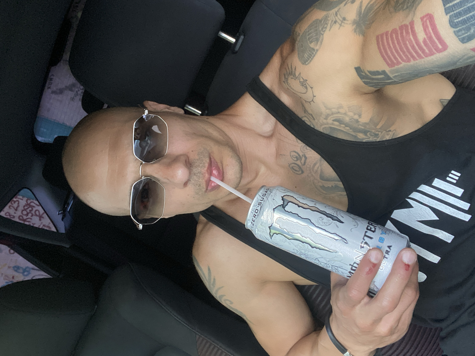 A bald man with sunglasses and tattoos is sitting in a car, holding a can of Monster Energy Ultra drink and drinking from it with a straw. The can is silver with green and white lettering. The man is wearing a black tank top. The car interior is visible, with dark seats and a headrest. In the background, there appears to be some colorful patterned fabric, possibly a blanket or clothing, and graffiti on a wall. The scene is shot from a selfie perspective. The location is not specified in the image or metadata.