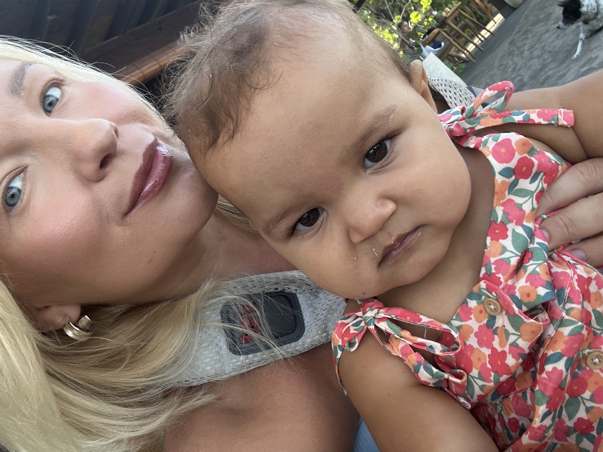 This close-up selfie features a blonde woman with blue eyes holding a baby with dark hair and brown eyes, set in an outdoor location in North Kuta, Indonesia. The woman is positioned on the left, looking directly at the camera with a gentle, slightly smiling expression. She has fair skin, wears light pinkish-brown lipstick, and a small silver earring. Part of her blonde hair frames her face. She is wearing a light-colored top or strap, possibly white or grey, with a black rectangular device, possibly a fitness tracker or camera mount, visible on her chest.
The baby, held close by the woman, is on the right, also looking directly at the camera with a neutral and curious expression. The baby has light brown skin, full cheeks, and appears to have some faint crumbs or residue around the mouth. The baby is wearing a colorful sleeveless dress adorned with a pattern of red, pink, orange, and green flowers on a white background, featuring tie-shoulders and small wooden buttons.
The scene suggests a bright, sunny daytime setting. The background is partially blurred but reveals an outdoor environment with hints of green foliage and wooden structures, possibly a patio or a garden area. In the upper right corner, a black and white dog is visible in the distance, walking on a paved surface, and some outdoor furniture (table and chairs) can also be faintly seen. No visible text is present in the image. The overall mood is intimate and warm.