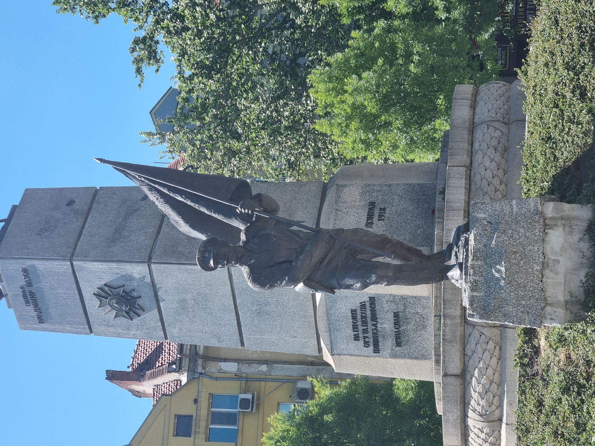 The image shows a statue in Pleven, Bulgaria, erected to honor soldiers of the 8th Pleven Infantry Division killed in battle at Doiran during World War I. The statue depicts a soldier with a flag. The monument consists of multiple stacked granite sections. The statue stands on a granite platform adorned with carvings. The sky is blue, and there is some greenery surrounding the monument and adjacent buildings. The buildings visible behind the statue appear to be residential structures.