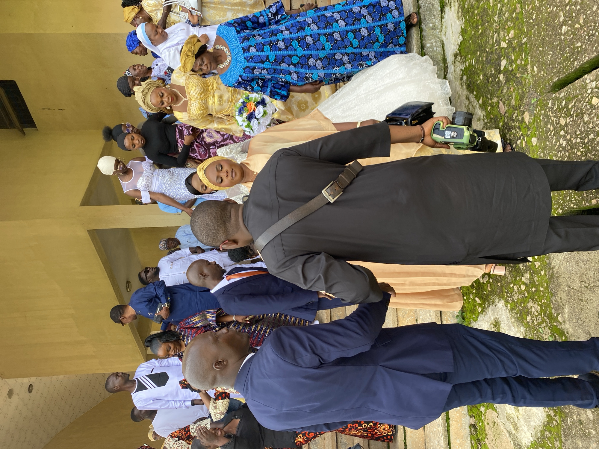 In Kuru Karama, Nigeria, a gathering of people, likely for a wedding or similar celebration, is captured outdoors. The image is oriented vertically, with the subjects appearing to be walking down a staircase or pathway. Several individuals are dressed in colorful, traditional attire, including women in vibrant blue and gold dresses and headscarves. One woman wears a white wedding dress, suggesting the event might be a wedding ceremony.
A man in a black suit is holding a green camera, possibly documenting the occasion. Other individuals are seen in suits and formal wear, indicating a celebratory atmosphere. The setting includes a building with a yellow facade and a moss-covered stone pathway or staircase, suggesting an outdoor location. The lighting indicates it is daytime. The people's expressions are mostly neutral or slightly blurred due to motion, but the overall impression is one of a formal gathering or celebration. No visible text is present in the image.