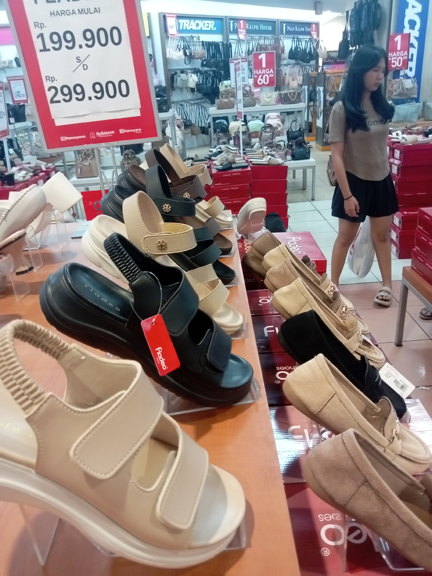 The image depicts a retail shoe store in Batam, Indonesia, likely a branch of Ramayana or Robinson department stores, as suggested by the signage. The focus is on a display of women's footwear, featuring sandals and loafers in neutral tones like beige and black. Some shoes are presented on stands, while others are stacked on shelves and display boxes. A large red sign prominently displays prices, indicating a starting price of Rp. 199.900 and another price of Rp. 299.900. In the background, a young woman with long dark hair, wearing a tan t-shirt and black shorts, is visible walking away from the camera, holding a white shopping bag. The store environment appears well-lit and organized, with shelves stocked with various shoe styles and handbags visible in the upper background. The overall impression is that of a typical department store shoe section.