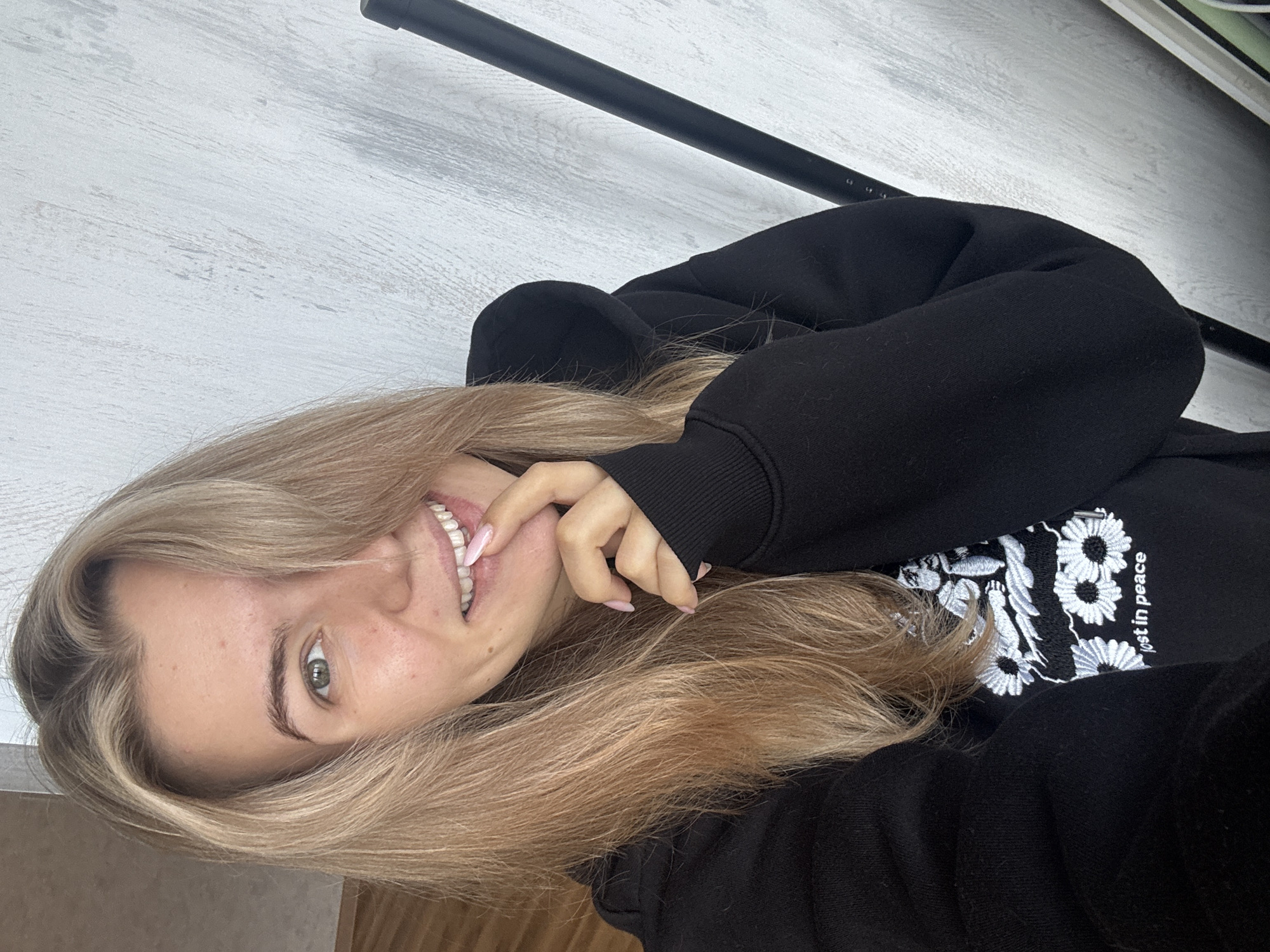 A young woman with long blonde hair is smiling and looking directly at the camera. She is wearing a black hoodie with a white floral design and the text "lost in peace" embroidered on it. She is holding her fingers near her mouth. The background includes a light-colored wall and a black rod or pipe running across the top of the frame. Location is unknown.