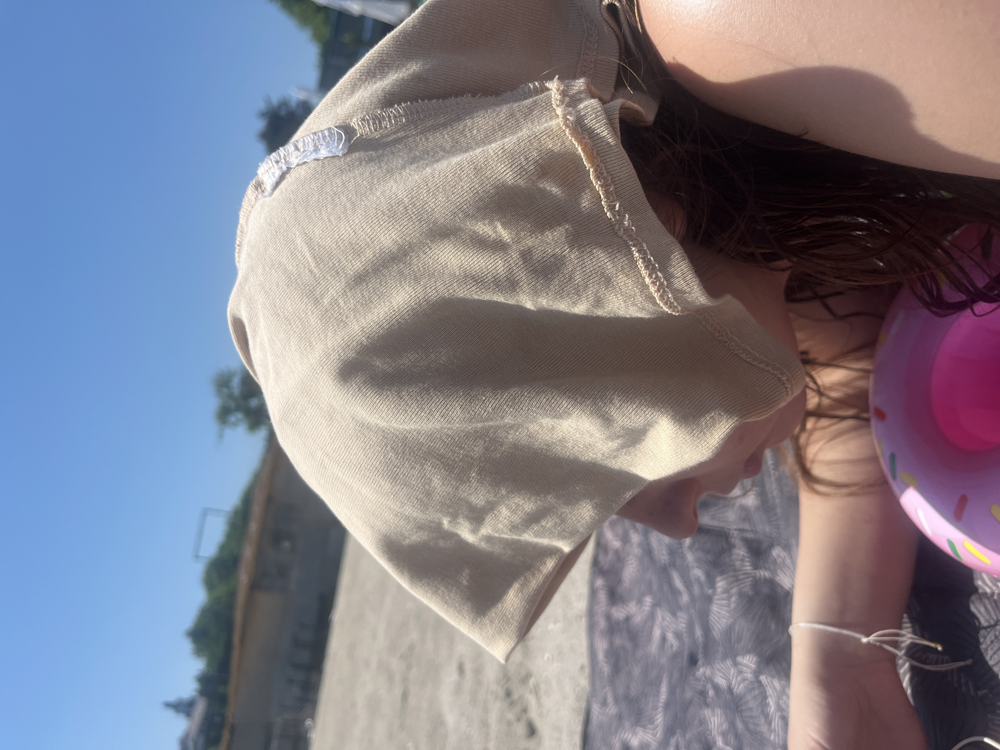 An overhead, rotated shot captures a person, likely a child or young individual, resting outdoors on a sunny day. The individual's head is largely covered by a light beige or tan hat or hood made of a ribbed fabric. Dark, wet hair spills out from beneath the hat, suggesting recent water activity. Only a small portion of their chin is visible. A white string bracelet is worn on their right wrist.
Beside the person, a pink inflatable ring, shaped like a donut with colorful sprinkles, is clearly visible. They are resting on a patterned blanket or towel, which lies on what appears to be sand or dirt. The background features a clear blue sky, with distant green trees and an indistinct structure on the horizon. The bright sunlight casts strong shadows on the hat and the person's exposed skin, indicating a clear, sunny day, likely mid-day or afternoon. A small white tag with faint, illegible text or a logo is attached to the rim of the hat. The overall scene suggests a relaxed leisure activity, possibly at a beach or by a body of water.