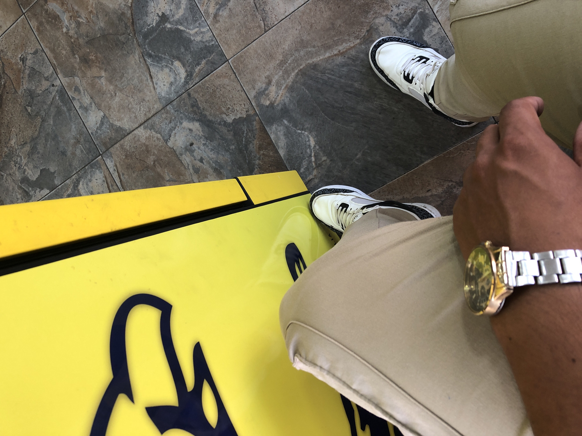 The image shows a person standing next to a bright yellow object with dark blue designs, possibly a sign or display. The person is wearing khaki pants and white sneakers with a black pattern on the sole. The person is also wearing a silver and gold wristwatch on their left wrist. The floor appears to be tiled with a gray, textured pattern. This image appears to be taken indoors, but the specific location in Juan Griego, Venezuela, cannot be determined from the visual information alone.
