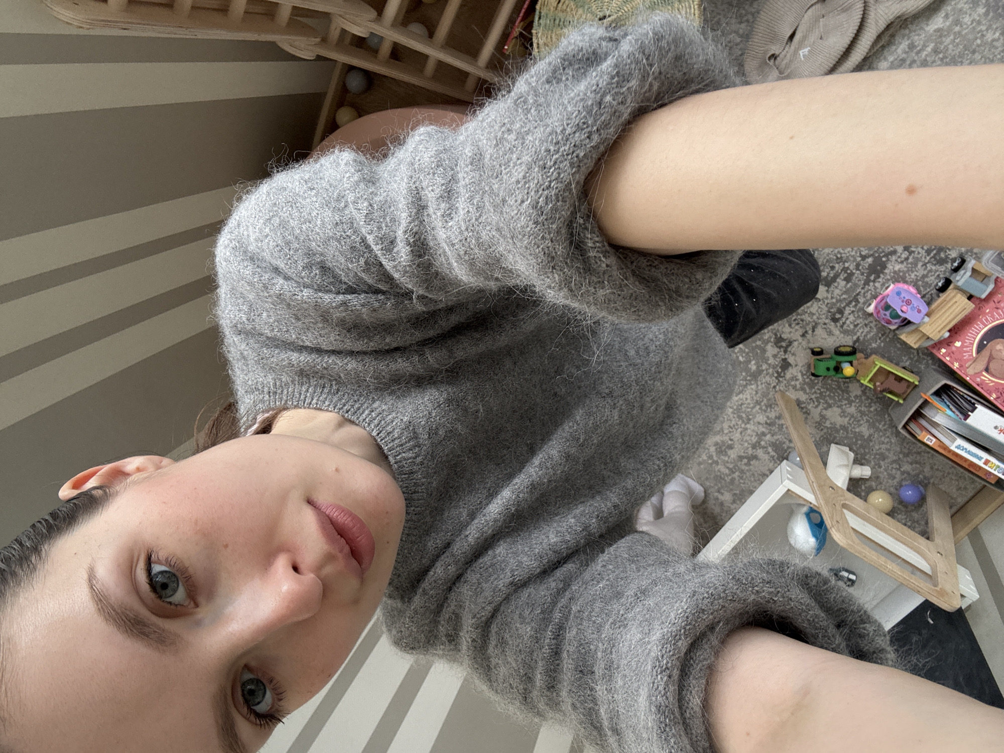 A young woman takes a selfie while wearing a grey sweater indoors. The background includes a striped wall, wooden furniture, and various children's toys and books scattered on the floor. The location is a domestic interior. There is no evidence of the image being taken in Moscow, Russia.