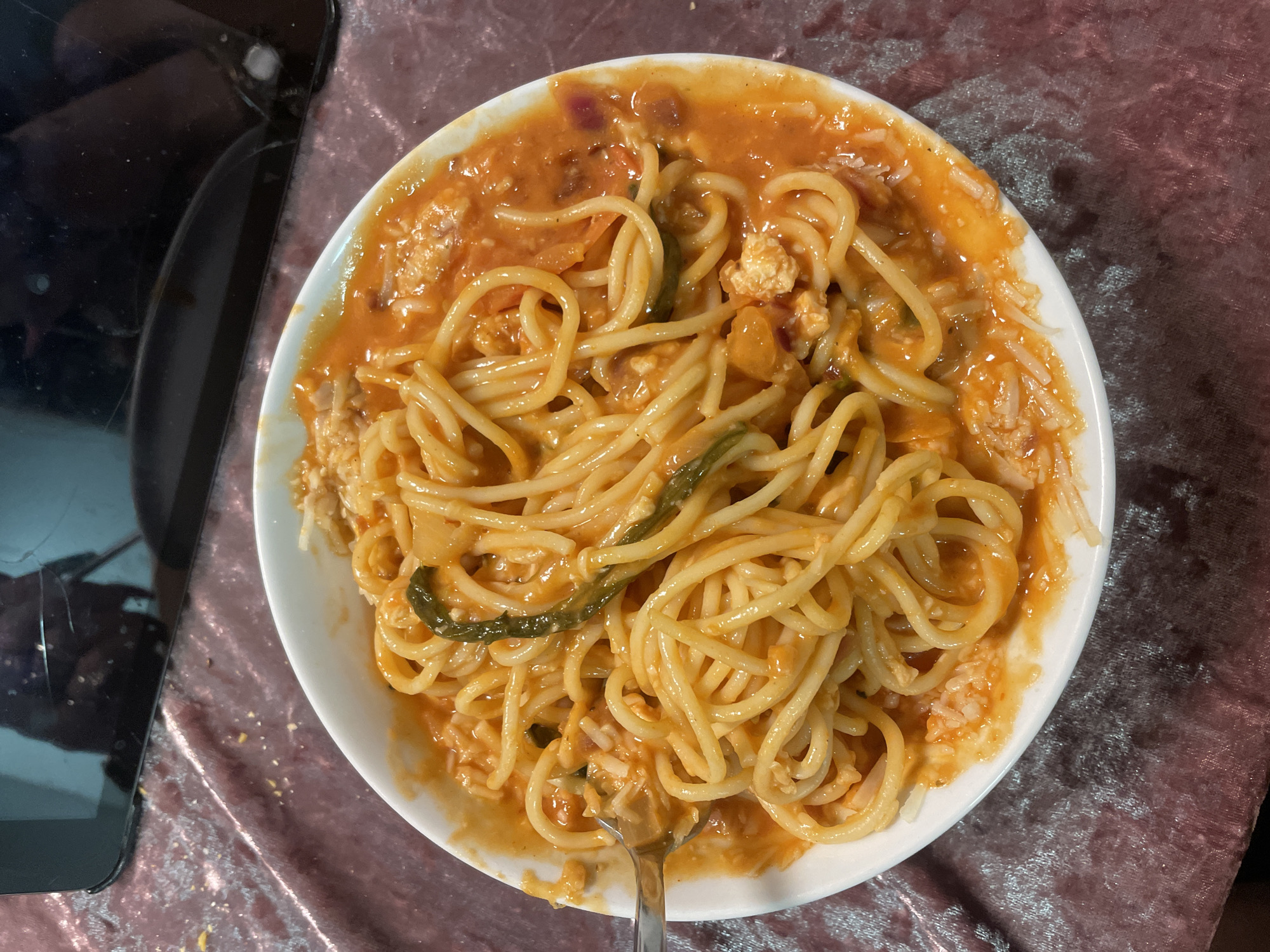 A top-down view of a white plate filled with spaghetti and a tomato-based sauce, garnished with what appears to be shredded cheese and some green vegetables. A fork is visible in the lower part of the plate. The plate is placed on a reflective surface that may be a table with a patterned tablecloth. A dark, possibly damaged, electronic device is partially visible on the left side of the image. No specific information suggests a location within Raanana, Occupied Palestine.