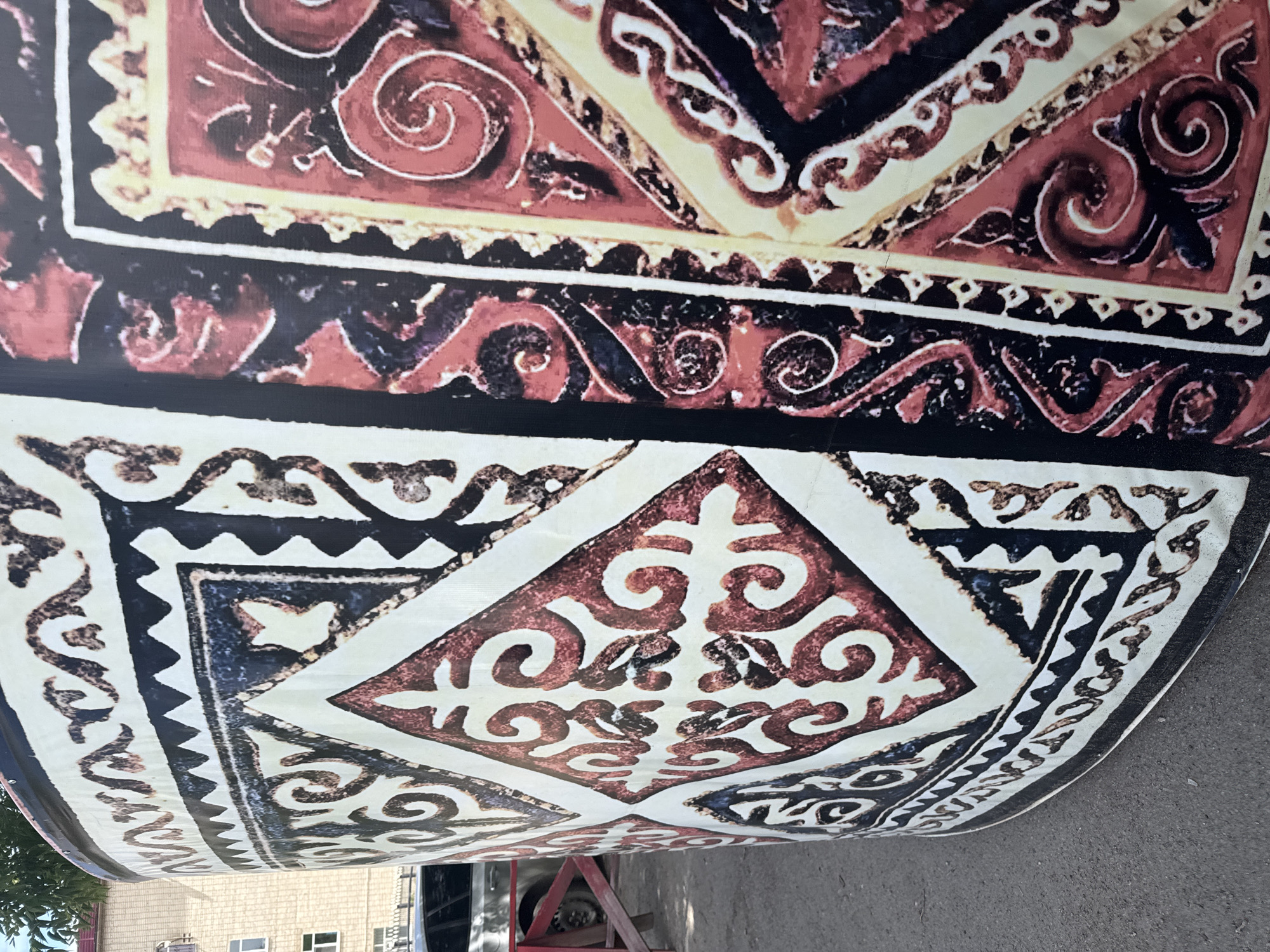 The image shows a close-up of a large, curved surface covered in a traditional pattern, likely a decorative wrap or mural on a structure. The pattern features geometric shapes and intricate swirling motifs in shades of red, black, and cream. The texture of the surface appears to be smooth. In the background, a brick building with windows is visible, along with a portion of a car and a red fence. The ground in the foreground is asphalt. This visual context suggests the object is located outdoors in an urban or semi-urban environment. Given the architectural style and the nature of the decorative pattern, it is plausible that this is a public art installation or architectural embellishment in Kokshetau, Kazakhstan, possibly representing local cultural heritage. No people are visible in the image, and no actions or interactions are taking place.