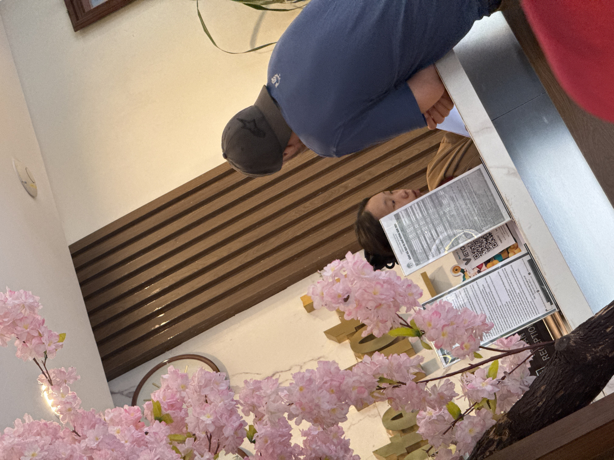 A rotated eye-level shot reveals a reception area in what could be a hotel or similar establishment in Binh Lu, Vietnam. A person wearing a dark cap and blue clothing is visible from the back. A reflection in what appears to be a glass or polished surface shows a woman's face. A sign indicating "RECEPTION" is seen, along with Vietnamese text that seems to be related to holiday pricing or services in 2025. There are decorative elements, including a clock and cherry blossom-like flowers. The backdrop is a beige wall with brown horizontal stripes.