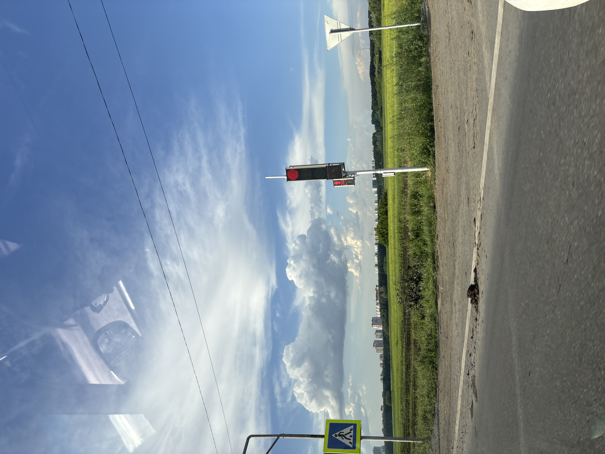The image shows a view of a road and a field under a blue sky with white clouds. On the left side of the road, a pedestrian crossing sign is visible. Further down the road, a traffic light with a red signal is present. In the background, there are buildings and a green field. The scene appears to be in a rural or suburban area. The location is identified as Elitnyi, Russia.