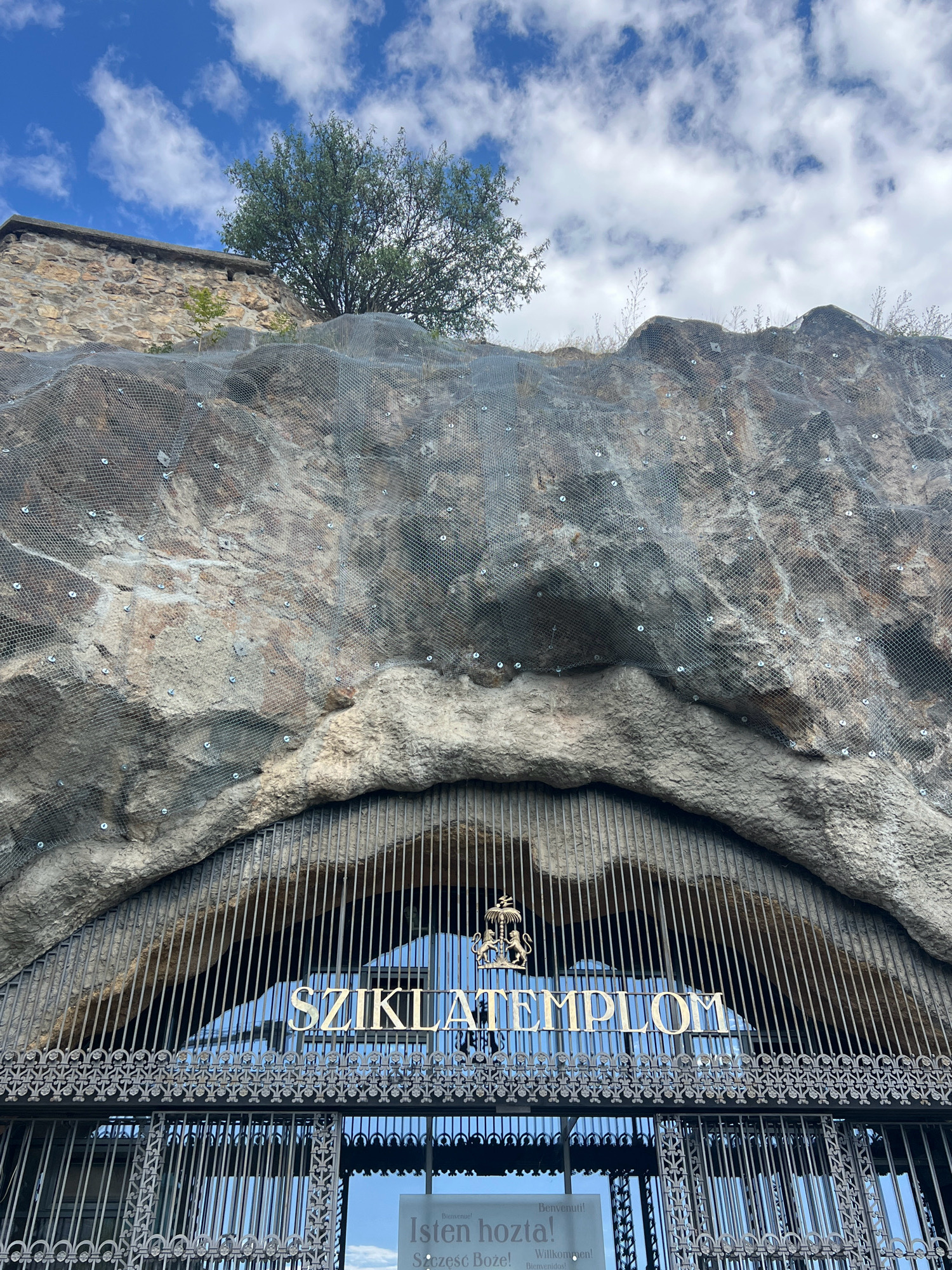 The image shows the entrance to the Sziklatemplom (Cave Church) in Budapest, Hungary, located at the Gellért Hill. The entrance is a large, arched opening in the rock face, covered by an ornate metal grate. Above the grate, gold lettering spells out "SZIKLATEMPLOM." The rock face itself is covered with a metal mesh, presumably for protection against rockfalls. A lush green tree grows from the top of the rock, and a stone structure, possibly part of the surrounding fortifications, is visible to the left. The sky above is blue with scattered white clouds. Below the main grate, a smaller gate is visible, and a sign with greetings in several languages, including "Bienvenue!" and "Willkommen!", is partially visible.
