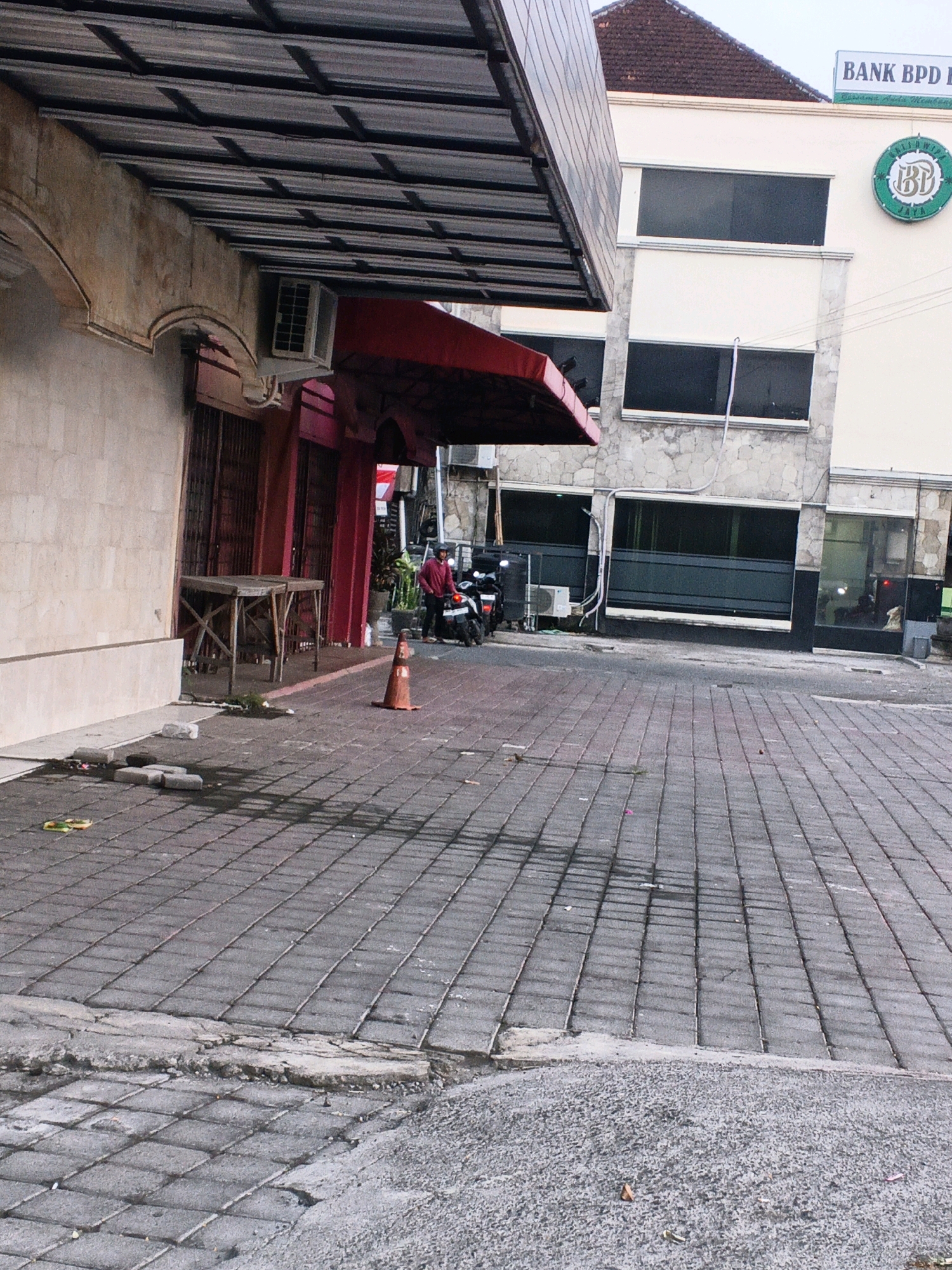 The image depicts a street scene in Kuta, Indonesia, likely in front of a commercial building. A white building with a stone-clad lower section and a concrete upper section is visible. The building has dark-tinted windows and a green circular logo with the letters "BP D JAYA" is mounted on the facade. A sign at the top of the building reads "BANK BPD". To the left, a section of a building with a beige textured wall and a metal awning is present. Under the awning, a red facade with closed metal shutters is visible. A simple wooden table stands in front of this building.
On the paved street in front of the buildings, a person in a red shirt is standing next to a parked motorcycle. An orange traffic cone is placed on the ground near the motorcycle. The street is paved with gray square interlocking bricks, and there are some areas of cracked concrete and debris on the ground. The overall lighting suggests it is daytime, possibly overcast or early morning/late afternoon. The environment appears to be a typical urban street with commercial establishments.