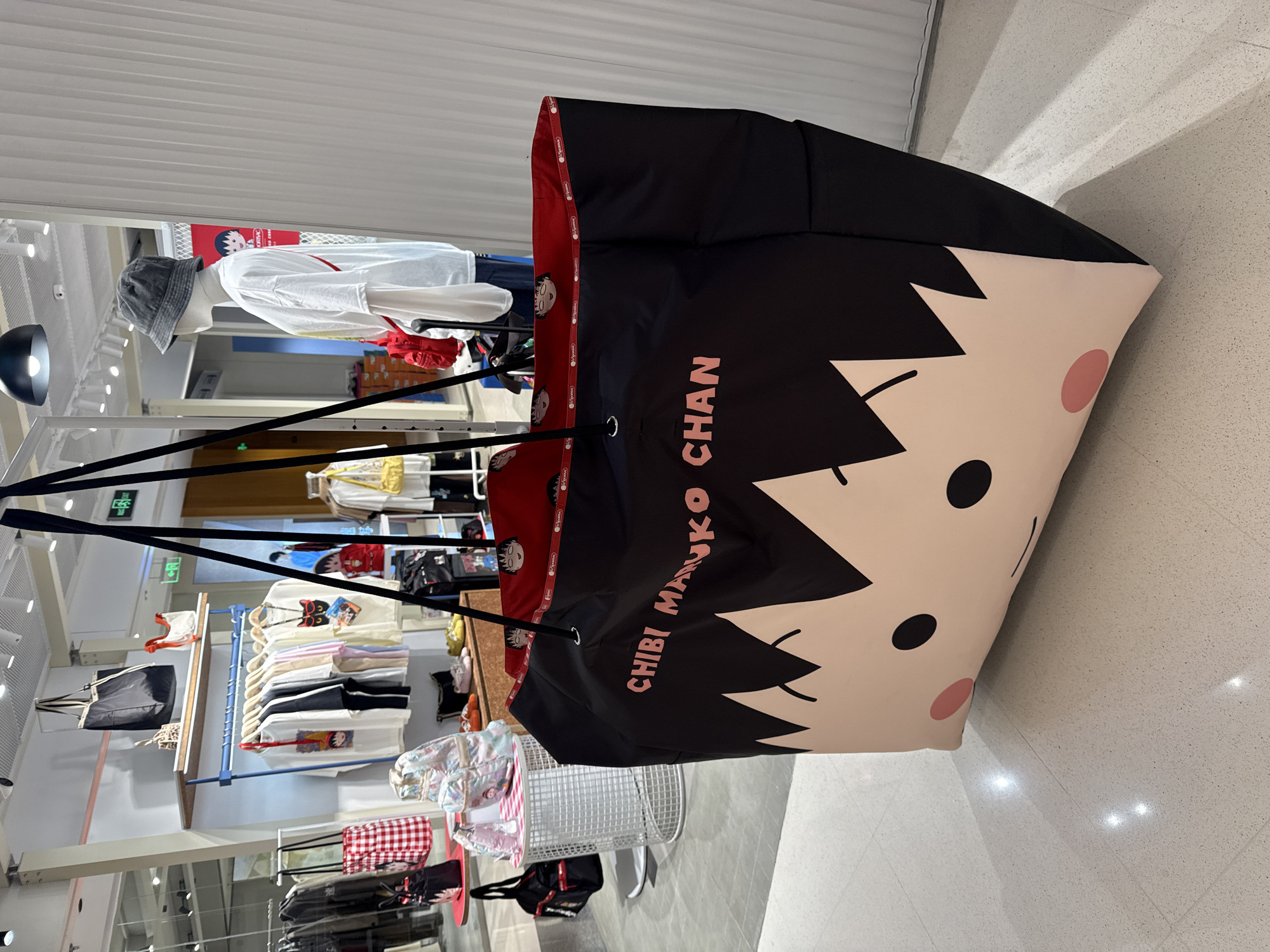 The image shows a large, black tote bag with a cartoon character's face and the words "CHIBI MARUKO CHAN" printed on it. The bag is hanging from black straps. In the background, a retail store displays clothing on racks and shelves, with mannequins dressed in apparel. The store appears to be a clothing boutique. The flooring is a light-colored speckled material. The scene is indoors and well-lit with overhead lighting. The image was taken in Shanghai, China.