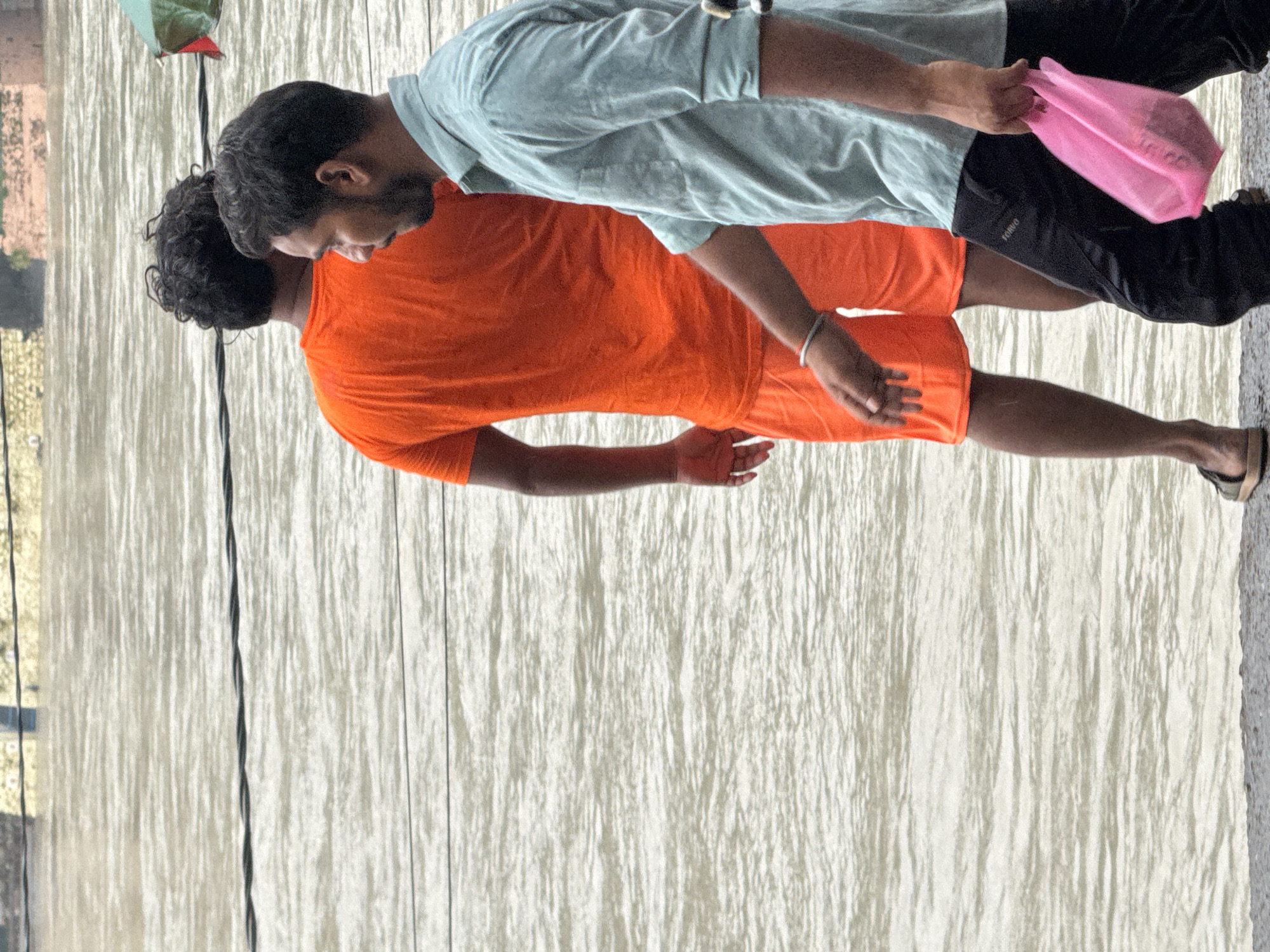 The image captures a section of the Ganges River in Rishikesh, India, with a low wall or embankment in the foreground. Two men are walking along the embankment, their backs mostly to the camera. The man in the foreground wears a light blue collared shirt and black pants, carrying a bright pink plastic bag in his right hand. He has a bracelet on his left wrist. The man behind him is dressed in an orange t-shirt and orange shorts, with dark, curly hair. The river water is a murky, light brown color, with visible ripples and some debris or electrical wires running across it in the background. The lighting suggests an overcast day.