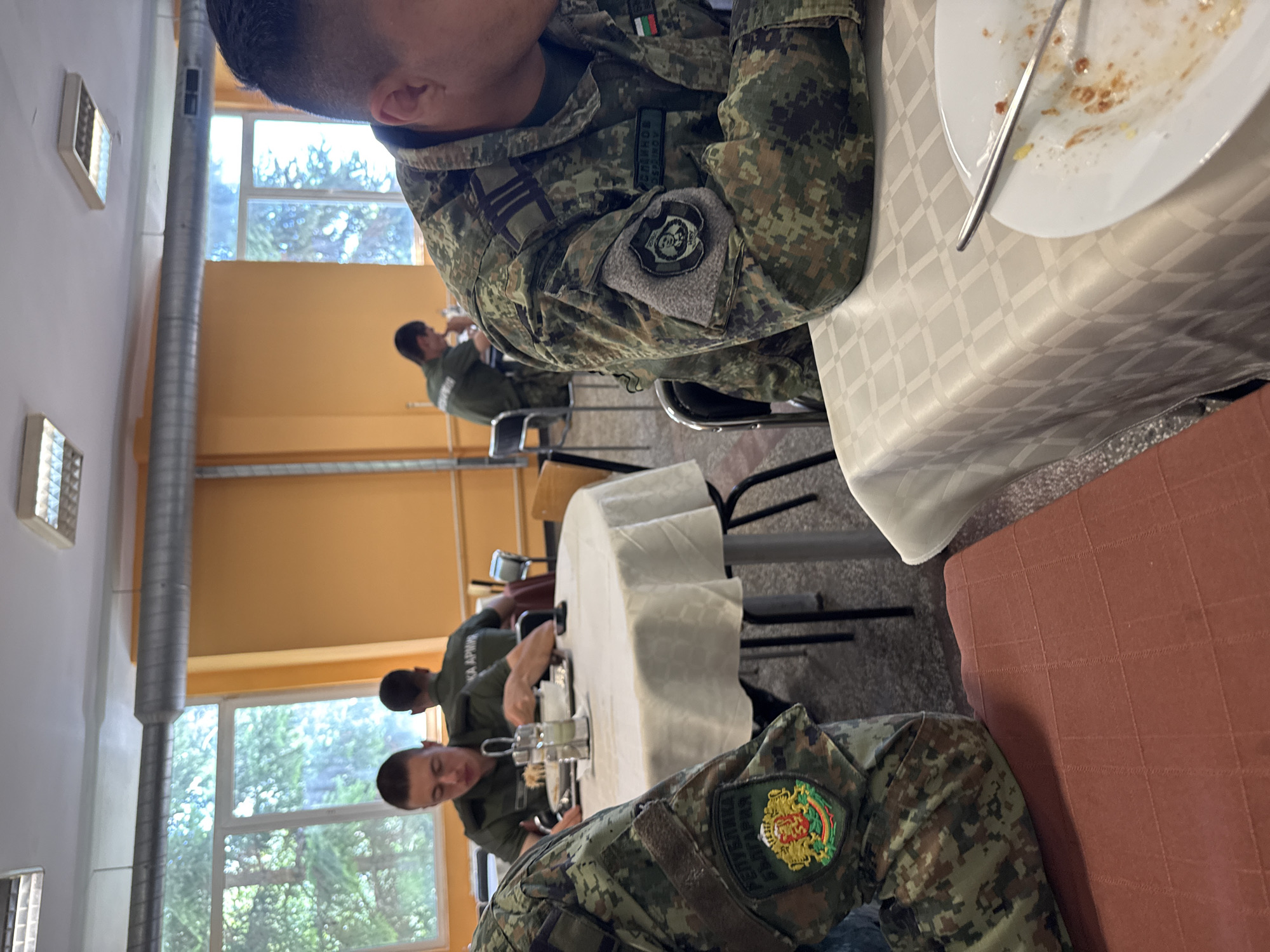 In a brightly lit canteen area, several individuals in military camouflage uniforms are seated at round tables covered with white tablecloths. In the foreground, a person in uniform with a patch showing a crest with lions and the text "РЕПУБЛИКА БЪЛГАРИЯ" on their left sleeve is partially visible. They are sitting at a table covered with a patterned reddish-brown cloth. To their right, another person in a similar uniform is seated at a table with a white tablecloth, with a partial view of their face and a plate with food remnants. Further in the background, two other individuals in military uniforms are seated at tables, with one person appearing to eat. The room has large windows with a view of trees outside, and orange-colored walls. Fluorescent lights are visible on the ceiling. The environment appears to be a mess hall or dining facility within a military base.