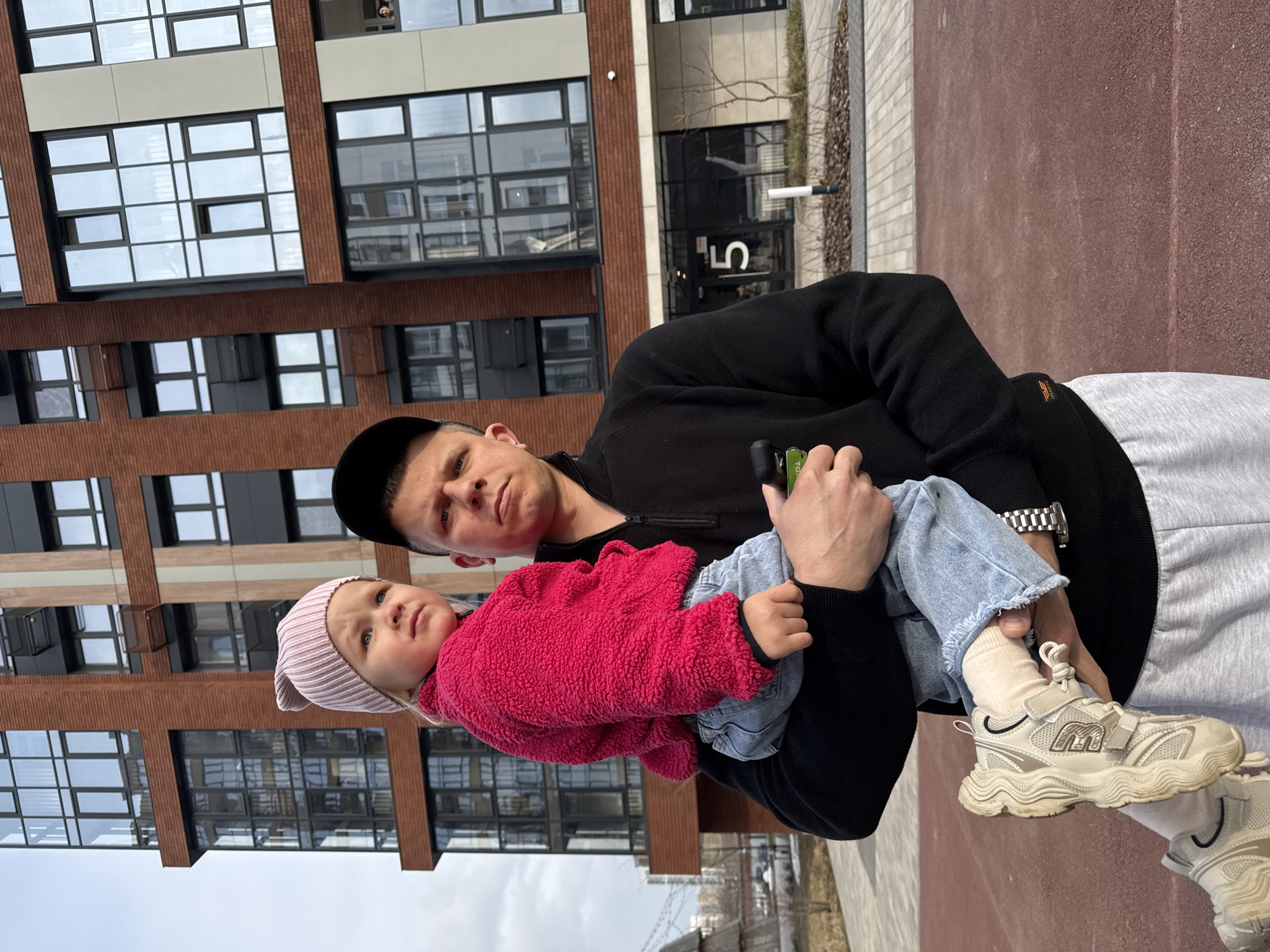 The image shows a man holding a young child in front of a multi-story building with brick facade and large windows. The man is wearing a black jacket, black cap, grey sweatpants and a silver wristwatch. The child is wearing a pink hat and a pink fleece jacket over a light blue jean. The background includes a modern building with a visible number "5" on it. The location is likely in a city, possibly Perm, Russia, given the architectural style and the visible Cyrillic-looking building numbers in the distance.