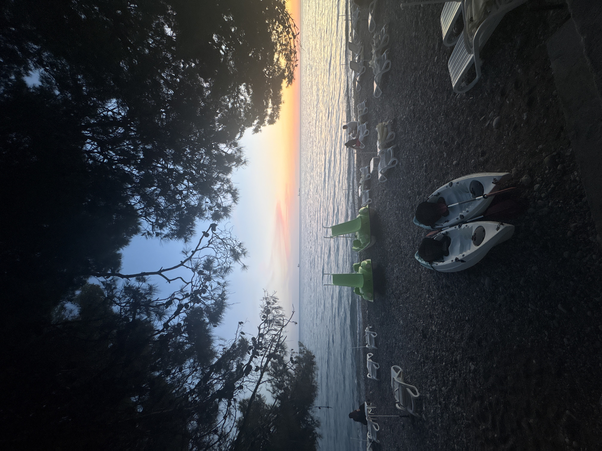 A dusky scene unfolds on a pebble beach in Bar, Montenegro, as the sun dips below the horizon. The foreground is dominated by dark, silhouetted pine branches arching over the frame, their needles creating a textured canopy. On the dark pebble ground, two kayaks, white with black seats and red paddles, rest side-by-side. Behind them, several white lounge chairs are neatly arranged, some occupied by figures, though their features are indistinct in the low light. Two bright green pedal boats are positioned further up the beach, closer to the water's edge. The gentle waves of the Adriatic Sea lap onto the shore, reflecting the soft hues of the sunset. The sky transitions from a pale blue overhead to pastel shades of pink and orange near the horizon, where a distant ship is visible. The overall atmosphere is serene and tranquil, capturing the quiet end of a day at the seaside.