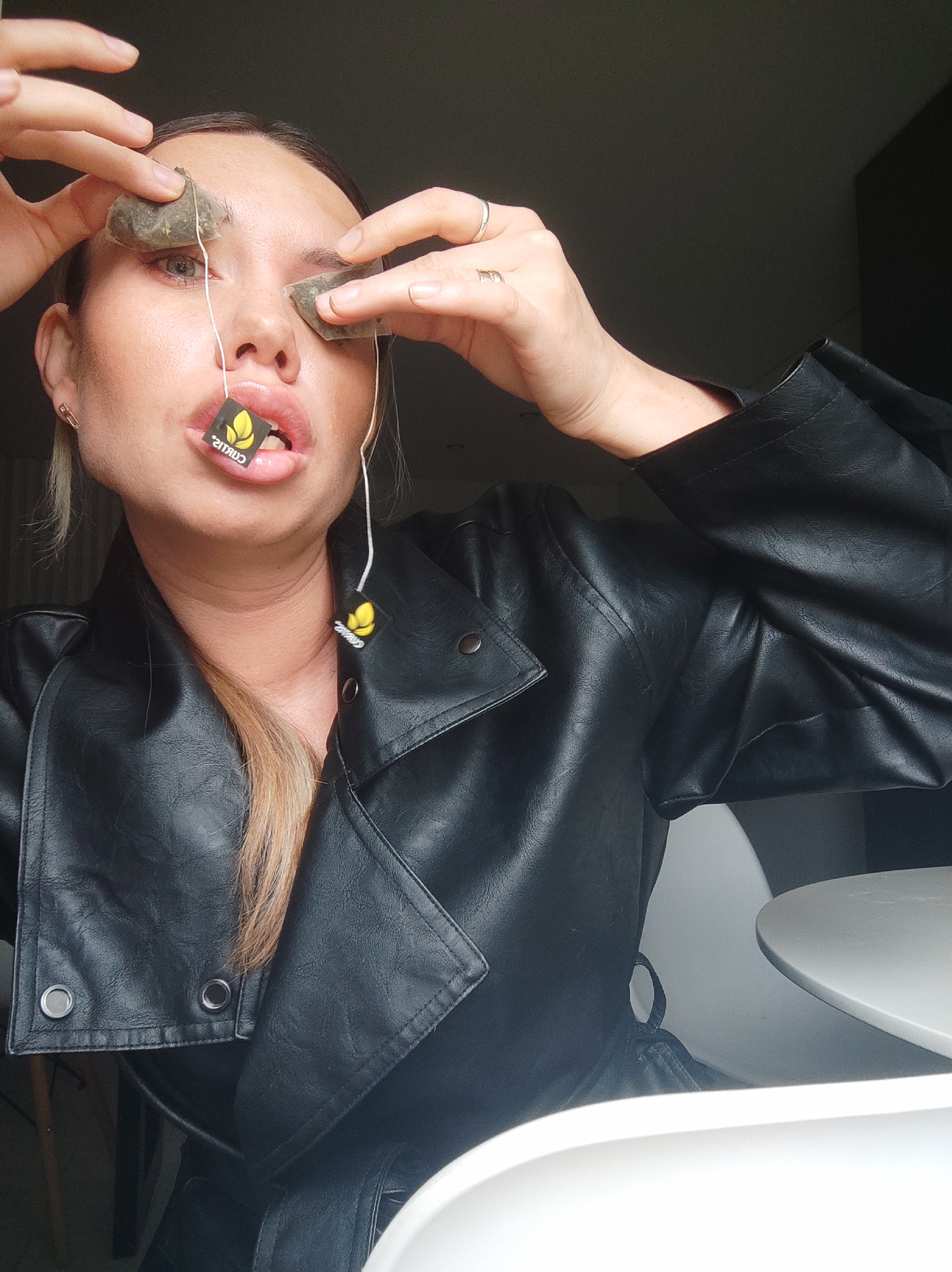 A woman is holding tea bags to her eyes, with another tea bag held in her mouth. She wears a black leather jacket. The background is a room with a chair and table visible. The location is unknown and cannot be identified as Votkinsk, Russia from the given image.