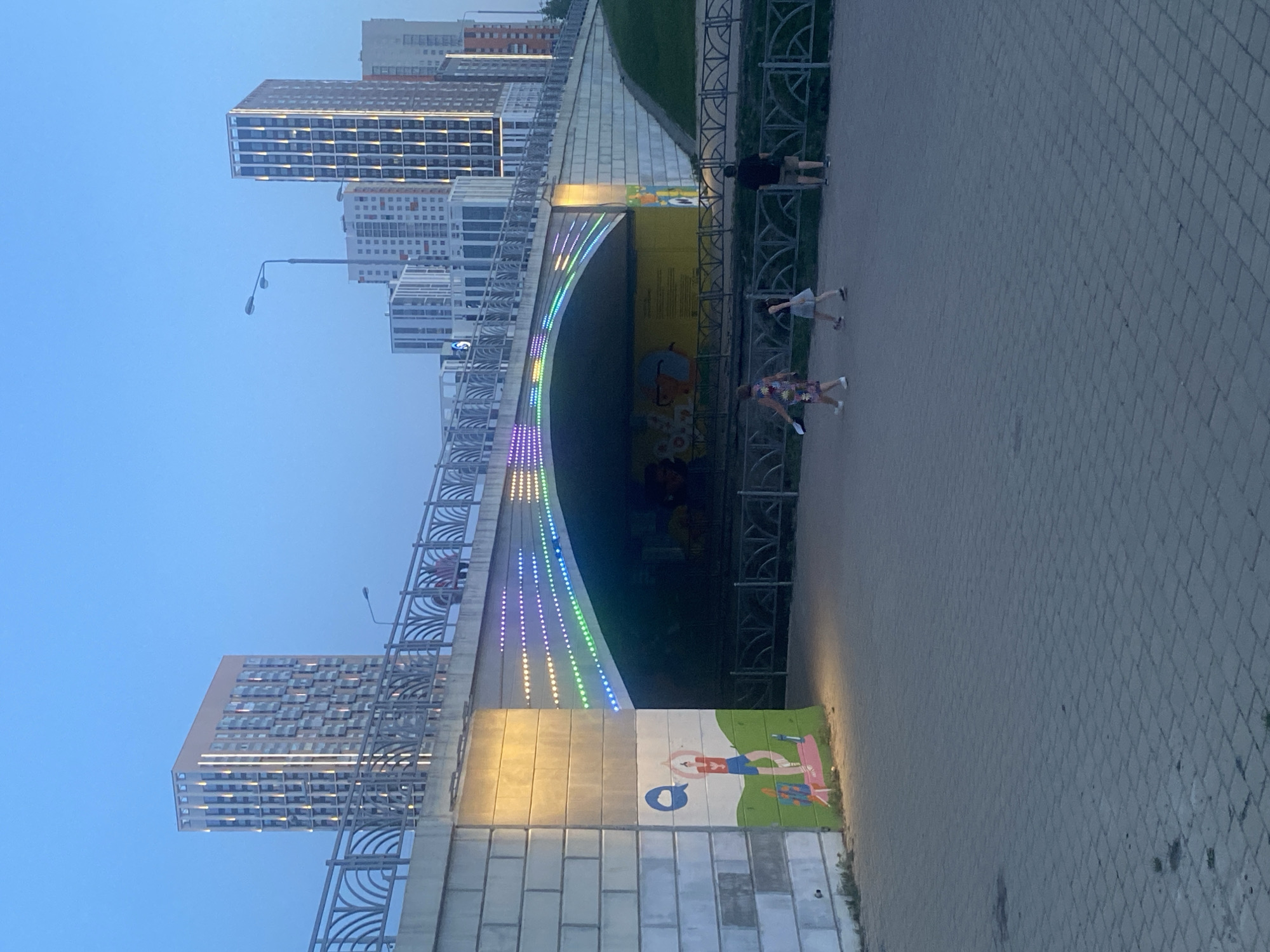 The image shows a section of the walkway near the pedestrian tunnel on Malysheva Street in Yekaterinburg, Russia. The pedestrian tunnel is underneath a bridge lined with buildings, and is illuminated with a line of multi-colored LED lights that run along the top edge, where it meets the bottom of the bridge. The walls are decorated with artwork. On the right side of the image, the pathway is paved with grey square bricks. There are three people walking on the path near the mouth of the tunnel. A metal fence separates the walkway from a grassy area. A tall residential building is in the background. The sky is blue.