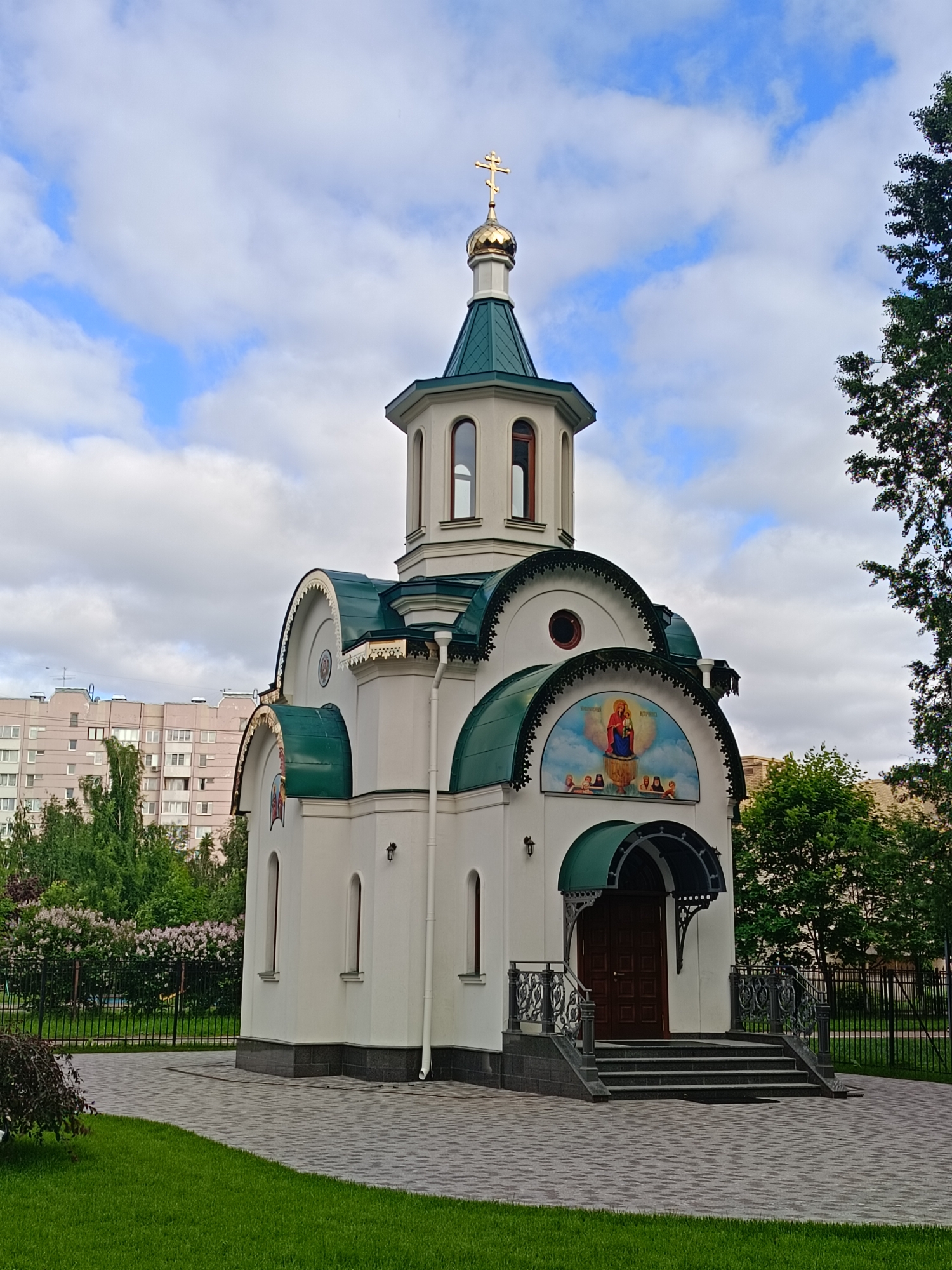 The image shows a small, white Orthodox chapel with a green roof and a golden dome topped with a cross. The chapel has arched windows and a decorated entrance. In front of the chapel, there is a paved walkway and a well-maintained lawn. To the left of the chapel, there is a residential building, and to the right, there are trees. The sky is cloudy with patches of blue. The chapel is identified as Chapel of the Intercession of the Most Holy Theotokos in Kolpino, Saint Petersburg, Russia. There are no people or animals visible in the image.
