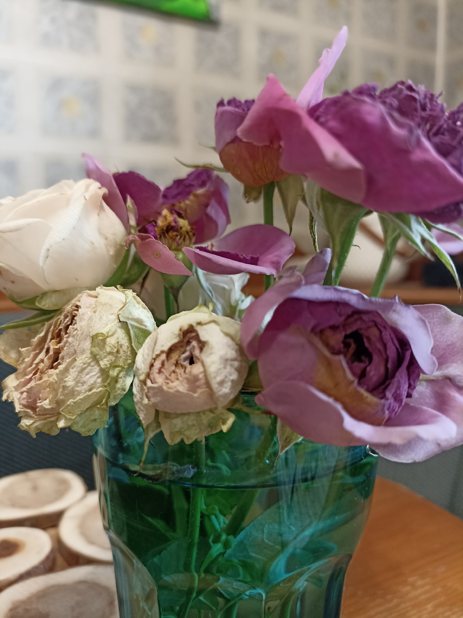 The image depicts a close-up, eye-level view of a bouquet of roses in a green glass vase. The roses are a mix of colors, including white, pale pink, and deep purple. Some of the roses appear wilted and dried, with brown and faded petals. The vase is filled with water, and the stems of the roses are visible submerged within. To the left of the vase, a stack of round, wooden slices is arranged on a wooden surface. The background is a wall with a subtle tiled pattern. The lighting suggests an indoor setting. There are no people or animals visible in the image. The image's composition focuses on the arrangement of the flowers and the texture of their petals. The overall scene appears to be a still life, likely set on a table or countertop. There is no information within the image that would indicate its location in Novosibirsk, Russia.