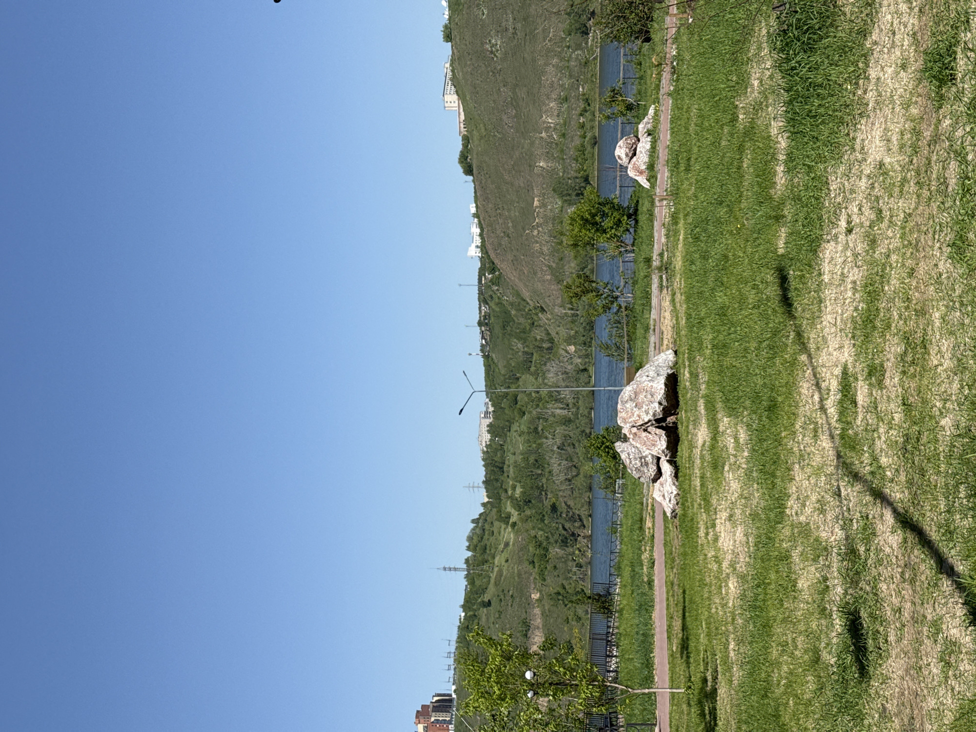 This image presents a serene outdoor landscape, likely a park or waterfront area in Krasnoyarsk, Russia. The scene unfolds under a clear, bright blue sky, indicating sunny weather.
In the foreground, a wide expanse of green grass with some drier patches stretches across the lower portion of the frame. A paved pathway, possibly for walking or cycling, runs horizontally through the grassy area. Several large, decorative rocks with mottled white and brown patterns are positioned near the pathway. Long, dark shadows are cast across the grass, suggesting it is daytime, possibly morning or late afternoon. Small green bushes and trees are scattered along the pathway and closer to the water's edge, and a single lamp post with a street light extends upwards from the left.
The midground features a calm body of water, likely a river, that spans horizontally across the image, reflecting the clear sky. A dark metal fence or railing separates the pathway from the water in some sections.
In the background, steep, verdant hills rise prominently, covered with dense green vegetation and some patches of lighter, drier earth. Several modern, light-colored buildings are visible on the slopes and crests of these hills, blending urban development with the natural landscape. Multiple tall power line towers or communication masts are also discernible along the ridge of the hills.
The scene is devoid of people, conveying a peaceful and tranquil atmosphere. No visible text is present in the image. The combination of a large river, green hills, and interspersed urban structures is consistent with the geographical characteristics of Krasnoyarsk.