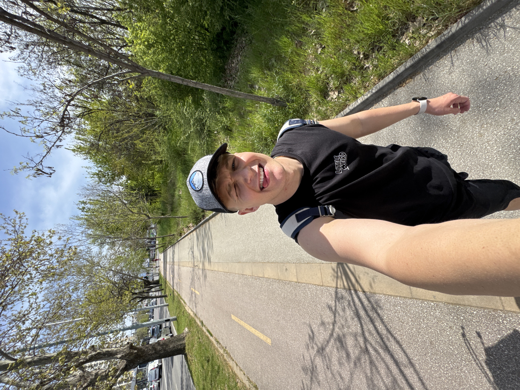 A sunny, outdoor selfie captured in Plovdiv, Bulgaria, shows a young male-presenting individual smiling playfully at the camera, with his tongue slightly extended. He wears a grey baseball cap with a blue and white emblem, a black t-shirt with the white text "LITTLE WEIRD" on the left chest, a white smartwatch on his left wrist, and a dark blue and white armband on his right arm.
The setting is a paved path, likely a walking or biking trail, stretching into the distance with a yellow dividing line. To the right of the path, there's a strip of green grass, and beyond that, dense foliage and trees, some with new, bright green leaves, indicating spring or early summer. To the left of the path, more trees line the edge, behind which a road with parked cars is visible. The sky above is clear and blue. The bright sunlight casts distinct shadows of trees and the person onto the path. The overall mood of the image is joyful and energetic, reflecting a pleasant outdoor activity on a clear day.