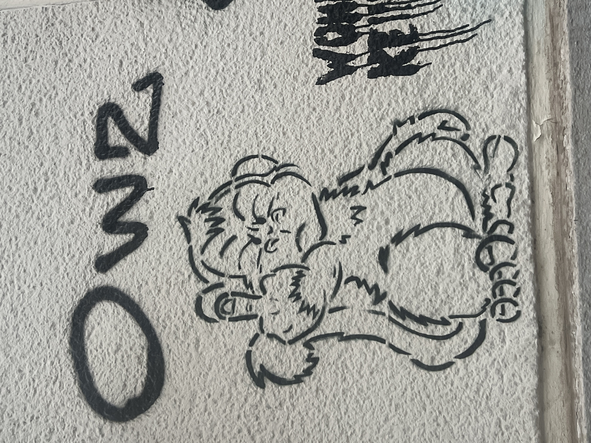 A black stencil of a cartoon monkey is spray-painted on a rough, white concrete wall. The monkey is depicted from the chest up, with its arms bent and hands held in front of its chest. It has a grumpy expression with furrowed brows and an open mouth showing sharp teeth. The monkey is giving the middle finger with its left hand. Above the monkey, the word "OWZ" is spray-painted in large, black block letters. To the right of the monkey, partially obscured, is another spray-painted word, possibly "WOAH" or "WOW", with some vertical drips of black paint below it. The lower part of the image shows a weathered concrete ledge with peeling paint. The graffiti is located on the exterior wall of the Urban Nation Museum in Berlin, Germany.