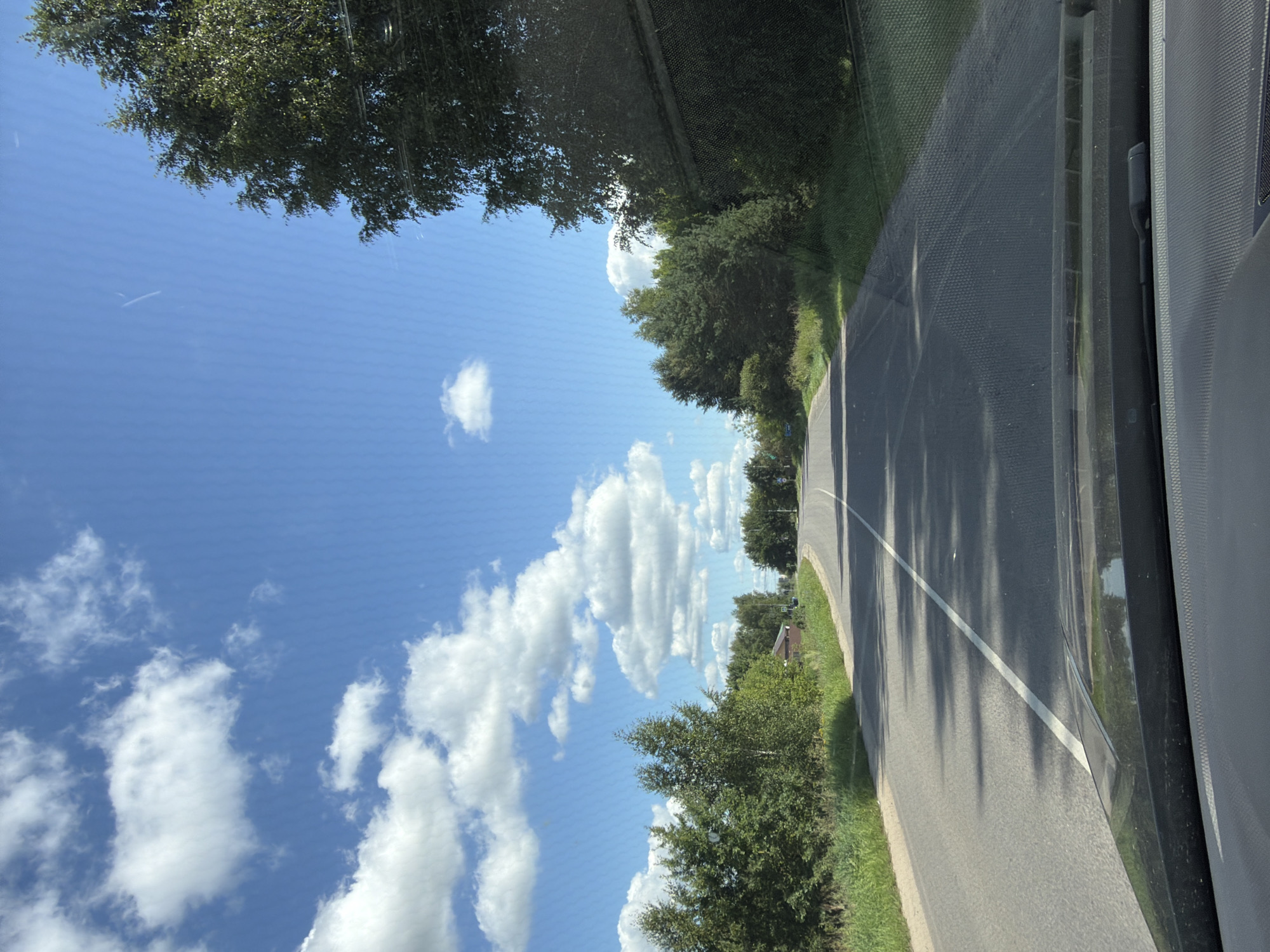 The image depicts a daytime scene of a paved road winding through a rural landscape. The road has a white dividing line and is bordered by green grass and trees on both sides. The sky is blue with scattered white clouds. The viewpoint is from inside a vehicle, showing the dashboard in the foreground. There are no people or animals visible in the image. The location is identified as a road in the village of Akseno-Butyrki, Russia. The environment suggests a sunny, possibly summer day.