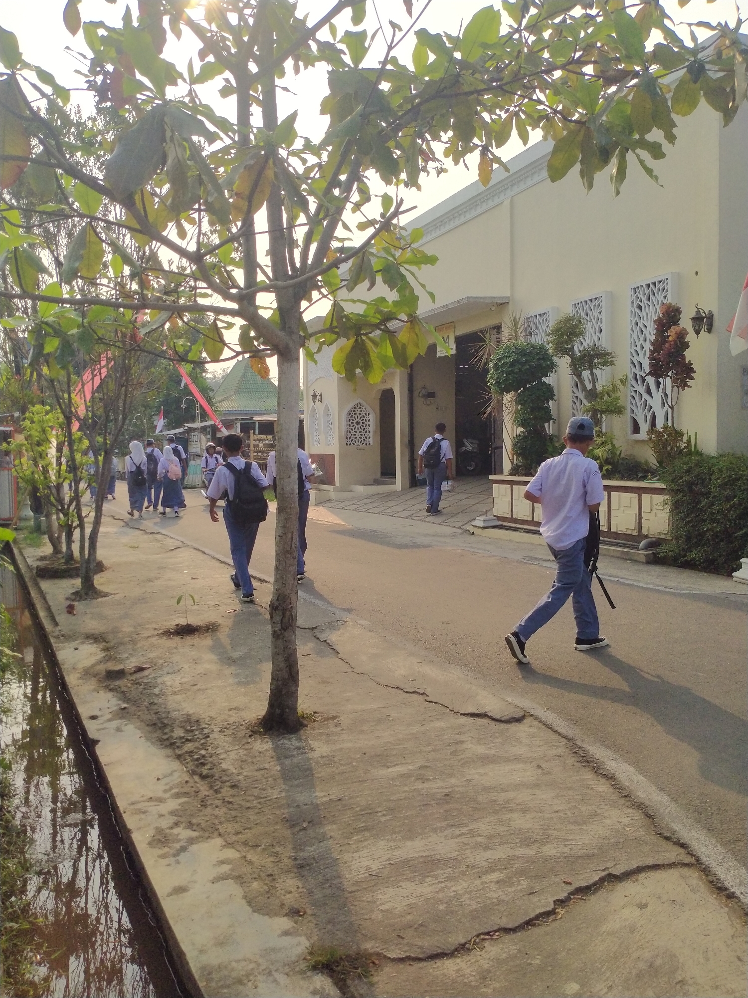 In the city of Begajah, Indonesia, a group of students in uniform walk along a paved path next to a road. The students, most of whom are wearing white shirts and dark pants or skirts, carry backpacks. The path appears to be on the left side of the road, bordered by a narrow canal filled with water and vegetation. Trees with green leaves line the path, casting shadows on the ground. In the background, a pale yellow building with decorative white windows stands beside the road. The sun is shining, creating bright light and shadows, indicating it is daytime.