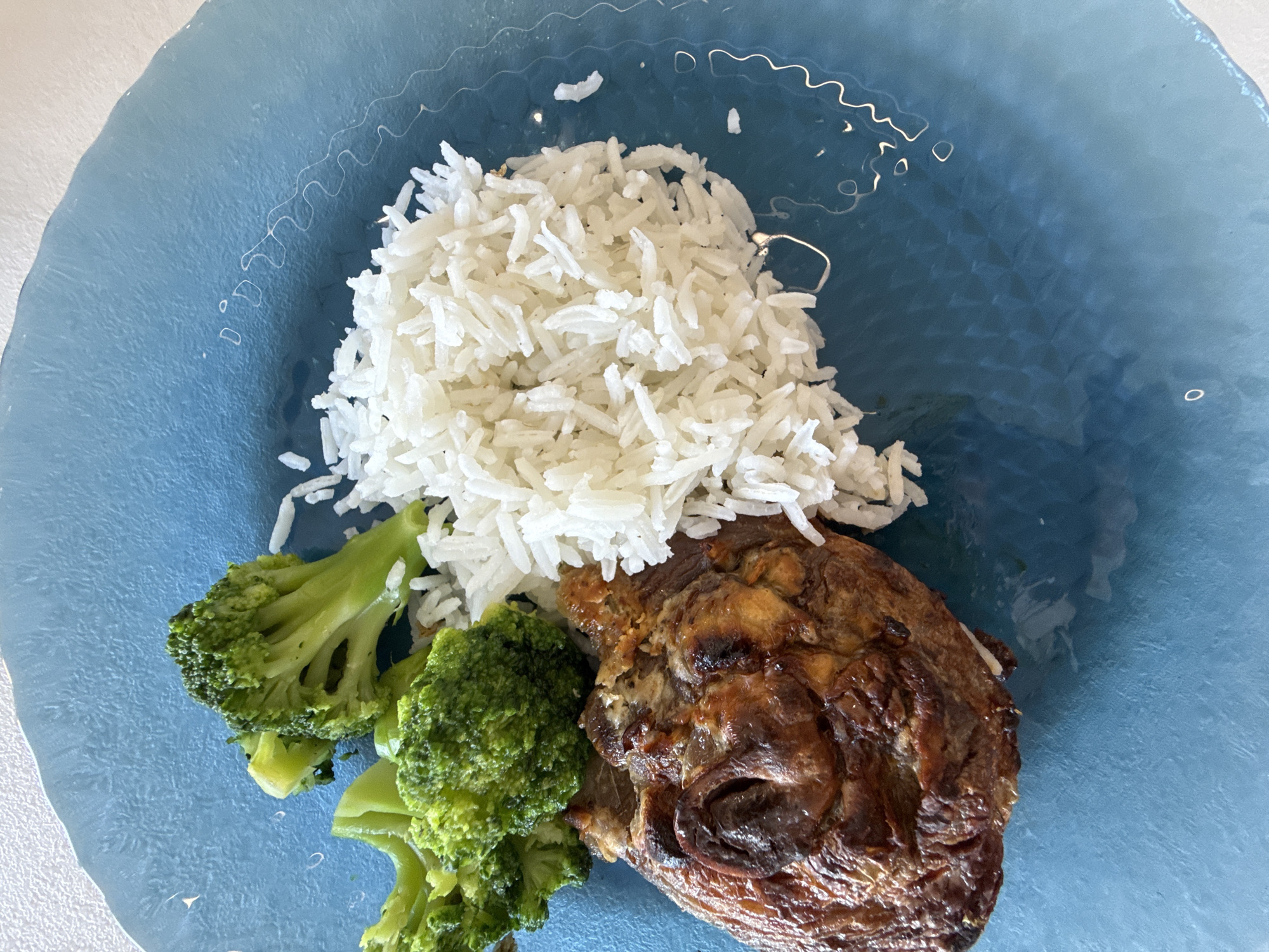 A blue plate holds a serving of white rice, broccoli florets, and a piece of browned meat. The image is taken in a close-up, overhead shot. There is no indication of the location being in Votkinsk, Russia.