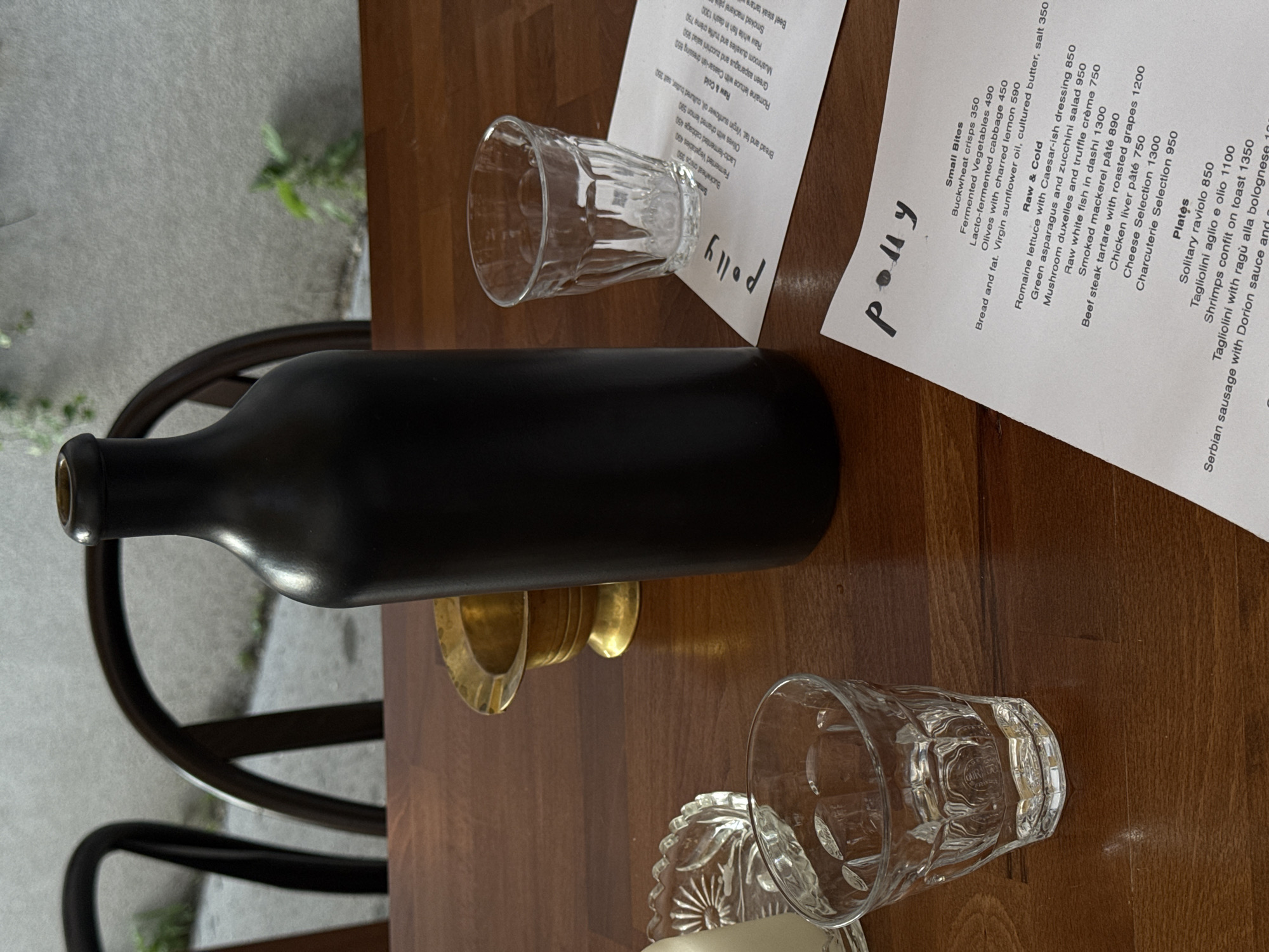 A dark matte bottle sits on a wooden table in a restaurant. The bottle is tall and has a narrow neck. Beside the bottle are two clear glasses, one empty and one with a small amount of liquid. A small brass ashtray is also on the table, along with a clear glass dish with a decorative edge. A menu is partially visible on the table, with the word "polly" written in cursive at the top. In the background, two dark wooden chairs are visible. The environment appears to be a restaurant in Belgrade, Serbia.