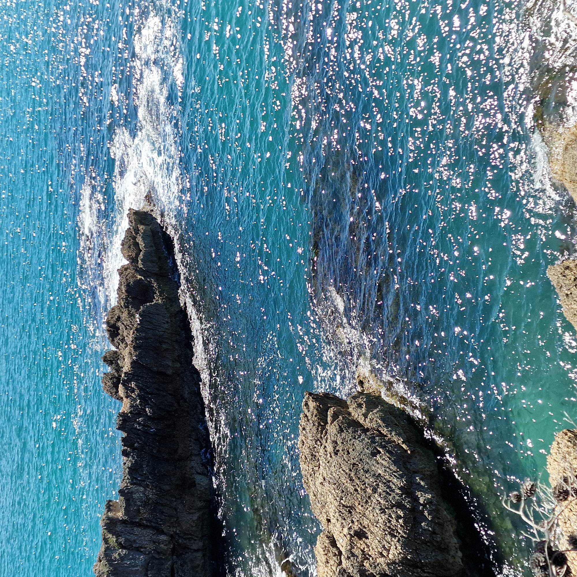 The image shows a rocky coastline next to the Adriatic Sea in Ulcinj - Ulqin, Montenegro. Jagged, dark gray rocks form a small peninsula. The sea is turquoise, with bright reflections scattered across the surface. Whitecaps are visible where the waves hit the rocks. There are tan-colored rocks on the right side of the image, showing some vegetation.