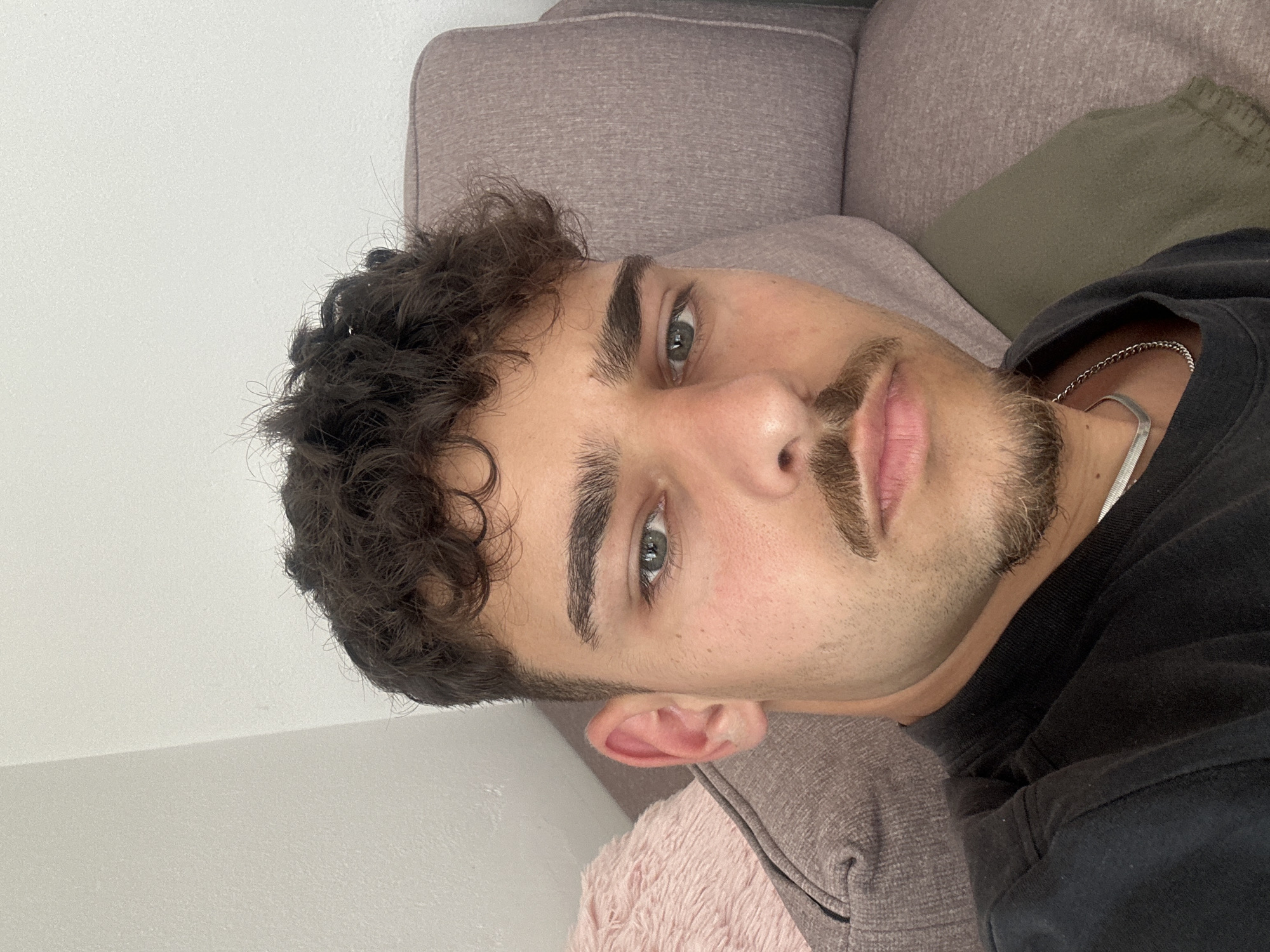 A young adult male with curly dark hair and green eyes is lying down on a light brown couch. He has a light complexion, defined eyebrows, a moustache, and a goatee. He is wearing a dark top and a silver chain necklace. A light pink pillow is visible near his left ear. The background includes a white wall. There is no evidence that the image was taken in the city of Troyan, Bulgaria.