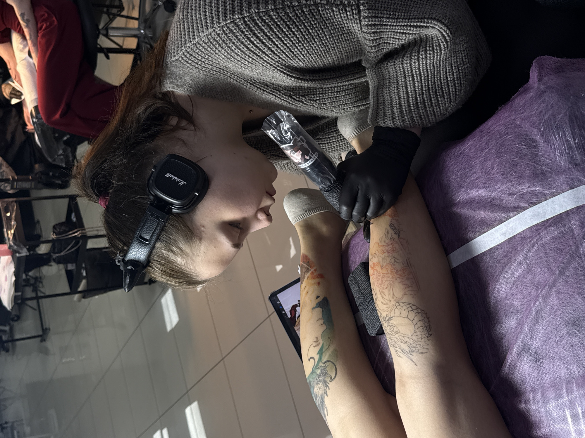 A person with dark hair and Marshall headphones is tattooing a design onto a person's leg. The tattoo artist is wearing a gray sweater and black gloves and holding a tattoo machine wrapped in plastic. The client's legs are resting on a purple, covered surface. Both legs have some tattoo design already present, and the artist appears to be working on one of these legs. A digital tablet rests near the design being worked on. The scene is inside a shop that likely specializes in tattoos, and the location is Rostov-on-Don, Russia.